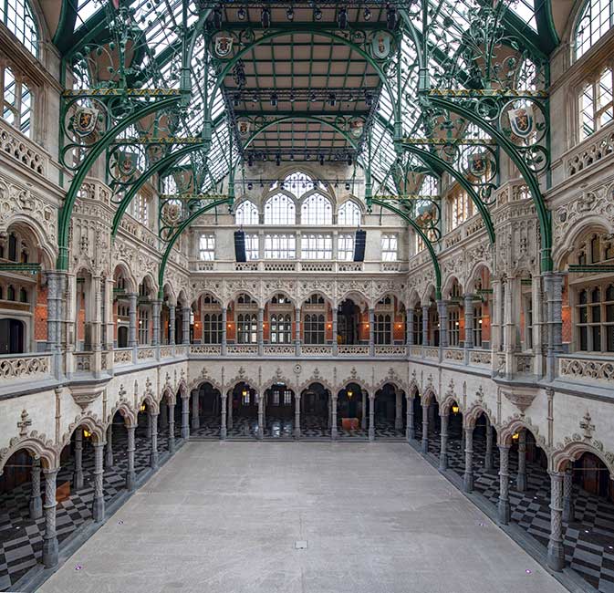 Gran vestíbulo interior neogótico de la Handelsbeurs Antwerpen con ornamentados arcos de hierro verde, techo de cristal y galerías de piedra con arcadas que rodean una gran planta abierta.