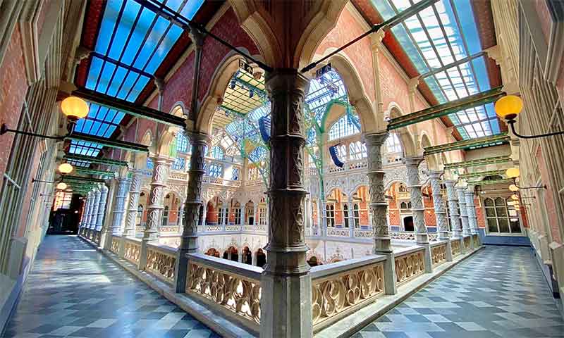 Vista de gran angular del vestíbulo interior neogótico con columnatas de piedra arqueadas, columnas ornamentadas, techo de cristal con paneles azules y suelo de baldosas a cuadros.