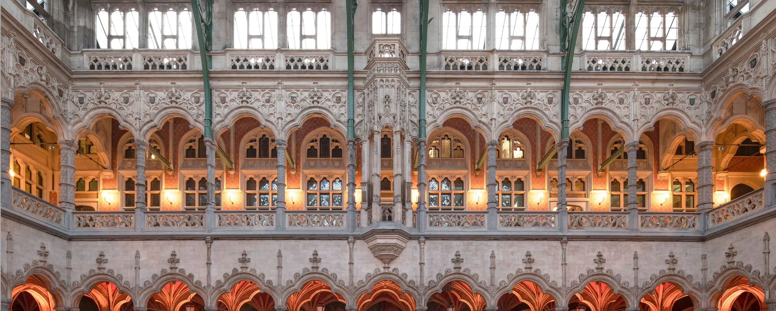 Vista panorámica amplia de las ornamentadas galerías interiores neogóticas con múltiples niveles de arcos de piedra y una cálida iluminación ámbar