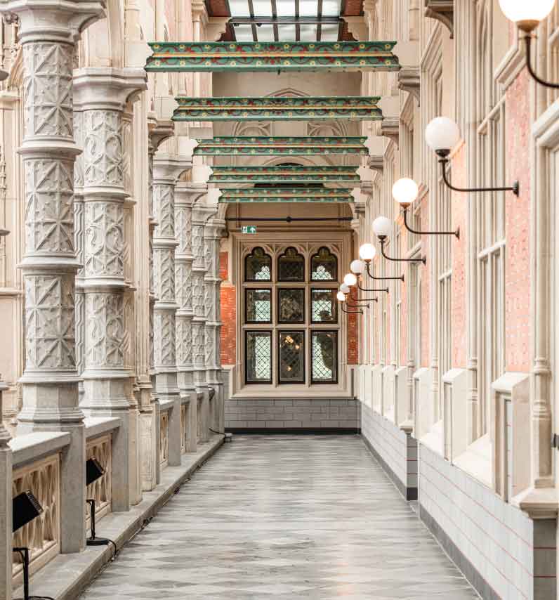 Un largo pasillo interior con columnas talladas blancas ornamentadas a la izquierda, vigas decorativas verdes en el techo y apliques de pared en forma de globo que conducen a una ventana arqueada.