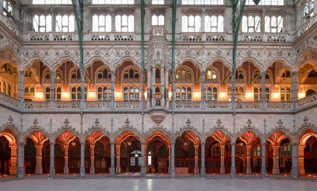 Vista interior panorámica de la Handelsbeurs Antwerpen neogótica que muestra tres niveles de ornamentadas columnatas arqueadas iluminadas con una cálida luz ámbar.