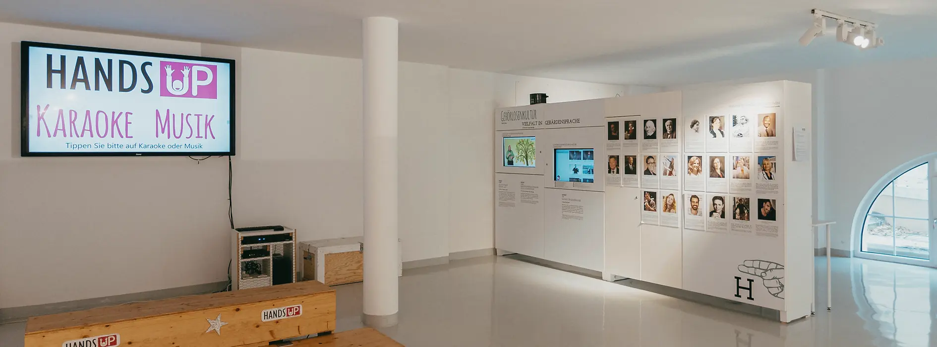 Espacio de exposición con una pantalla que muestra 'HANDS UP KARAOKE MUSIK' y una pared de retratos