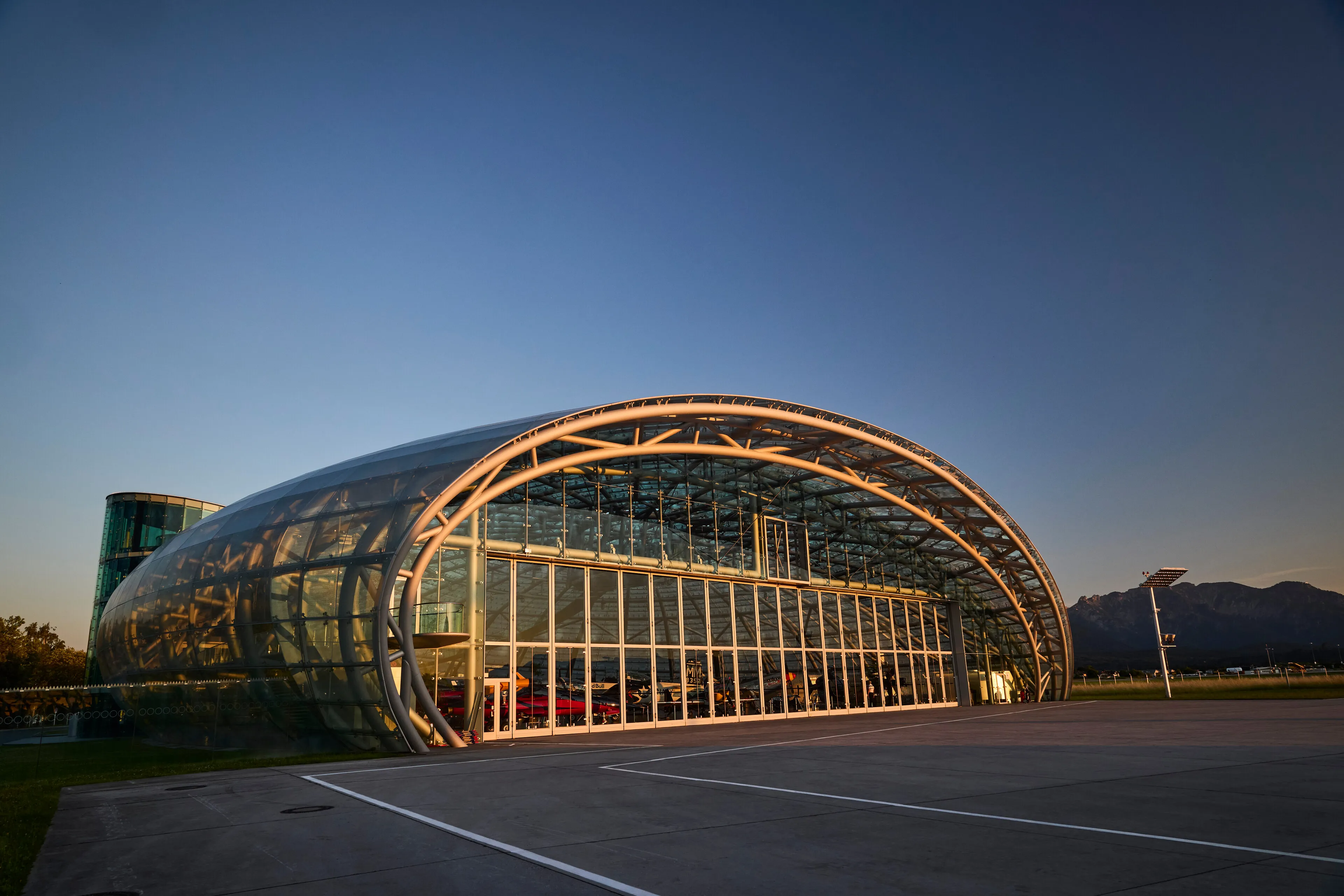 Gran edificio tipo hangar de cristal y acero con forma de arco, iluminado al anochecer con montañas al fondo y un aparcamiento en primer plano