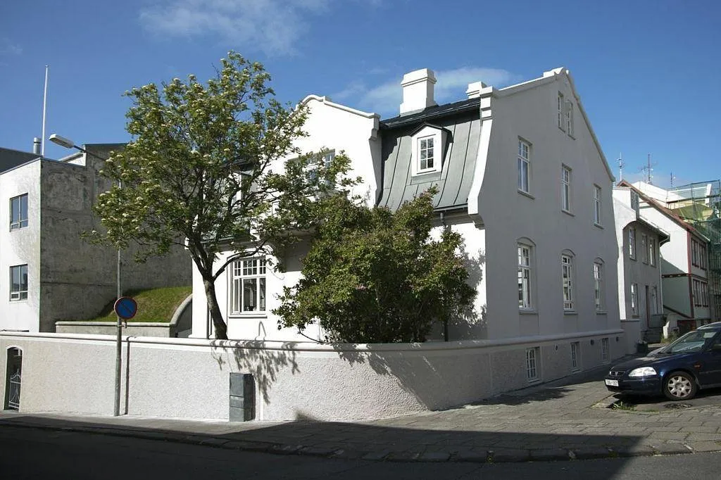 Edificio blanco de varias plantas con tejado abuhardillado oscuro, un árbol grande delante, un muro perimetral blanco bajo y un coche aparcado en una calle empedrada bajo un cielo azul