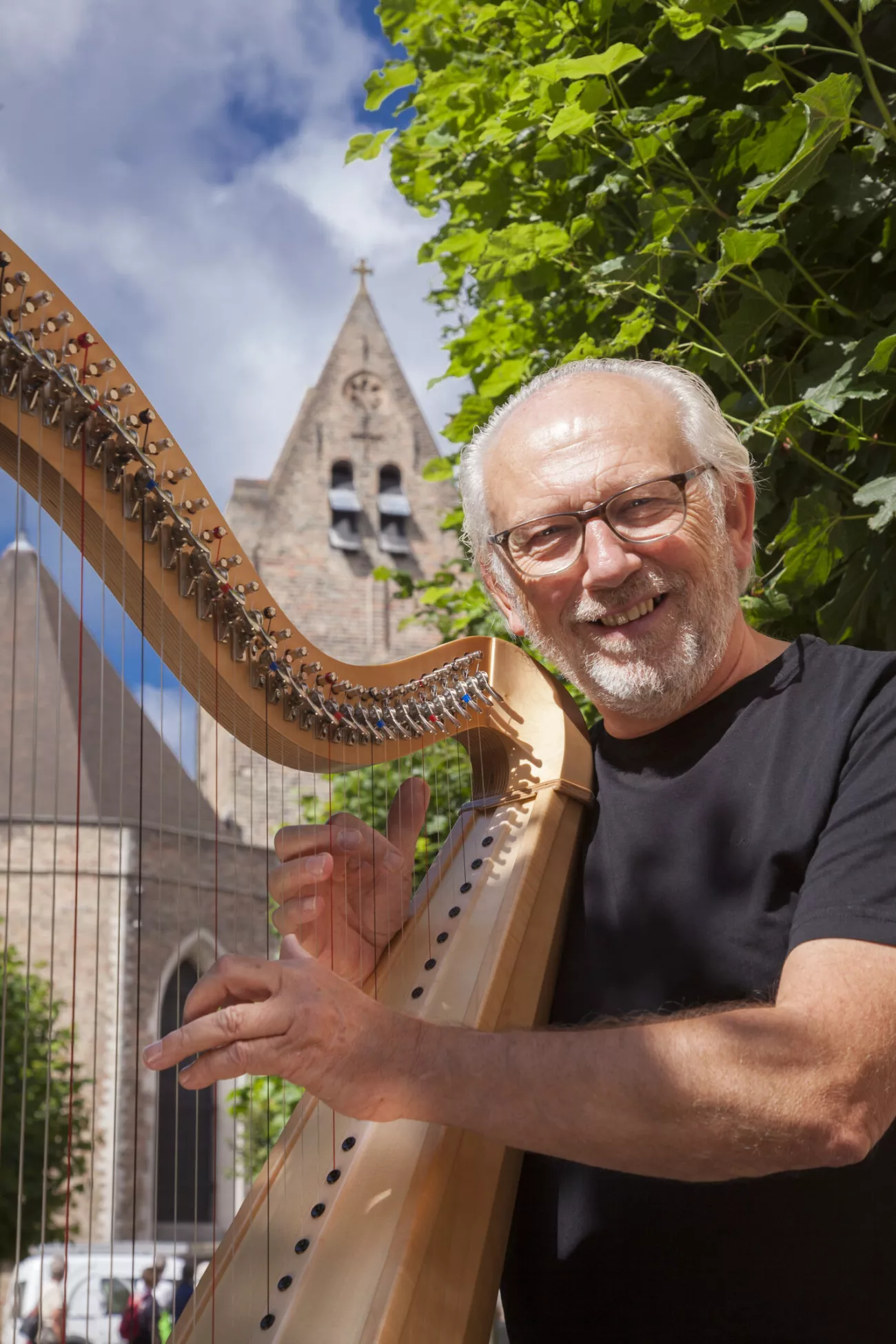 Un hombre mayor sonriente con gafas y barba blanca sosteniendo un arpa de madera al aire libre, con una torre de iglesia histórica y hojas verdes de fondo.