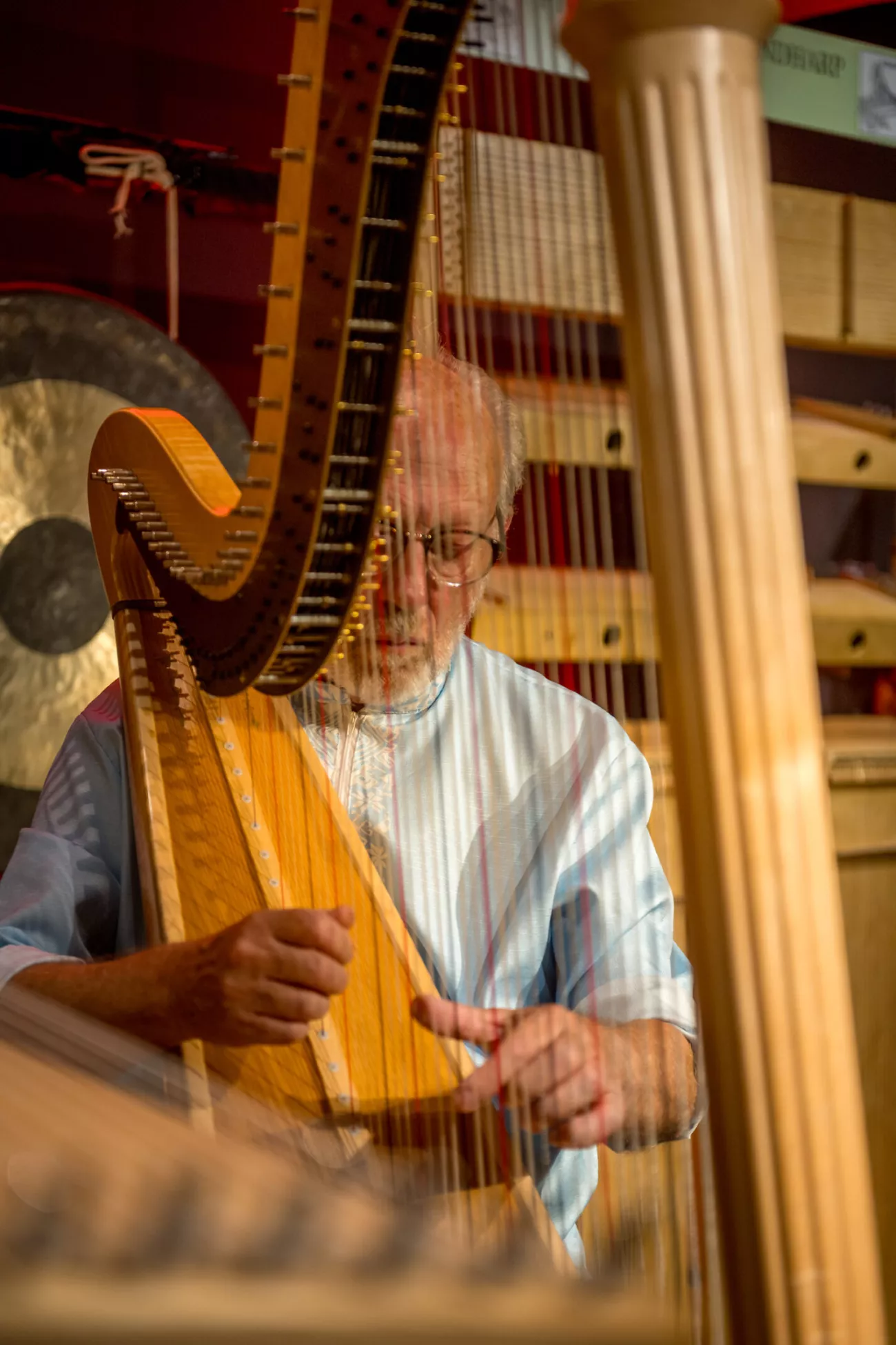 Un hombre mayor con gafas y barba blanca tocando el arpa, visto a través de las cuerdas de otra arpa en primer plano, con un gran gong visible al fondo.