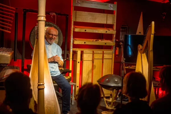 Un hombre con gafas y camisa azul claro se sienta entre varios arpas e instrumentos musicales de madera en una habitación de paredes rojas, dirigiéndose a una audiencia cuyas siluetas son visibles en primer plano.