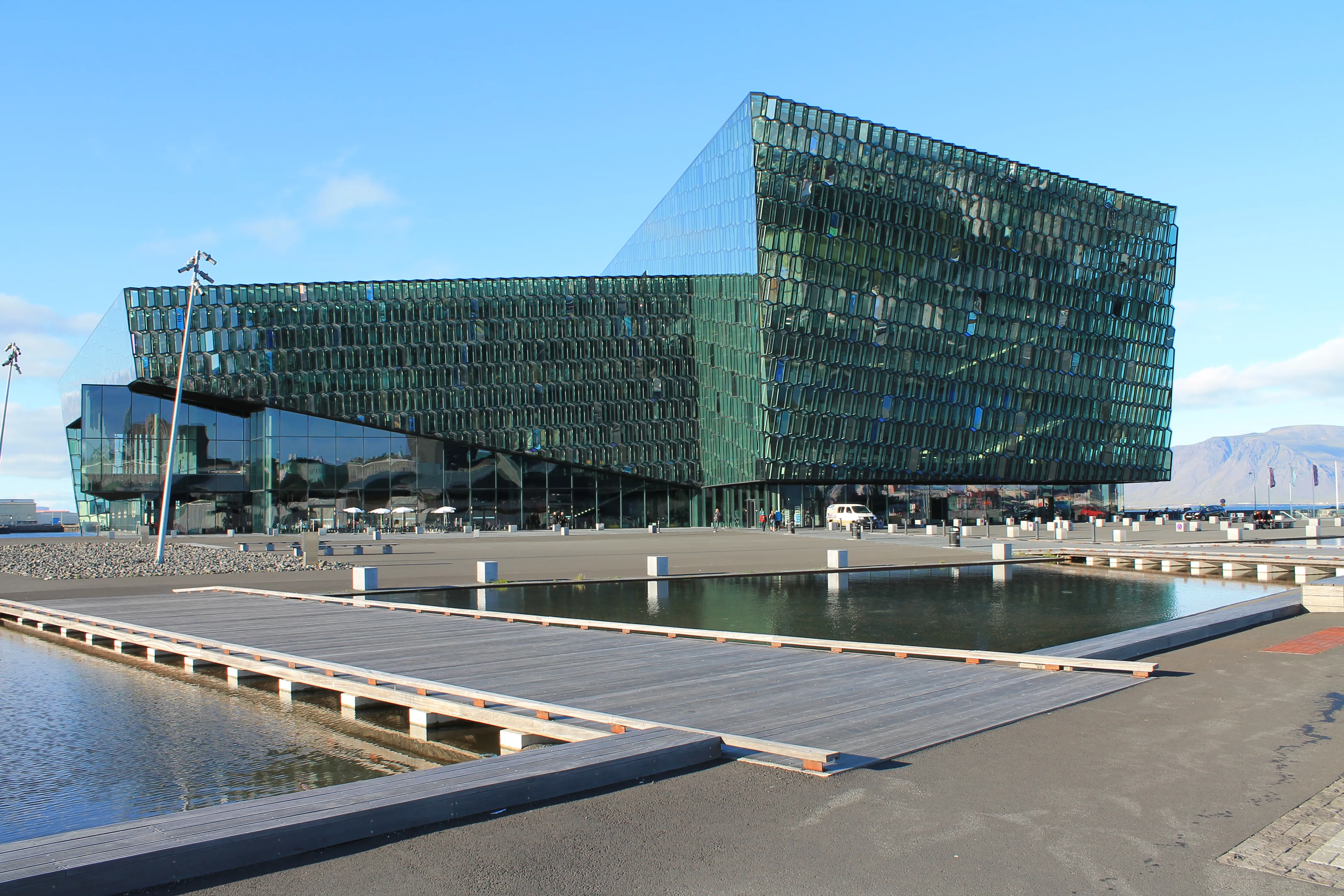 Vista exterior del Harpa Concert Hall que destaca por su gran fachada geométrica de cristal verde, un paseo de madera y un estanque en primer plano bajo un cielo azul.