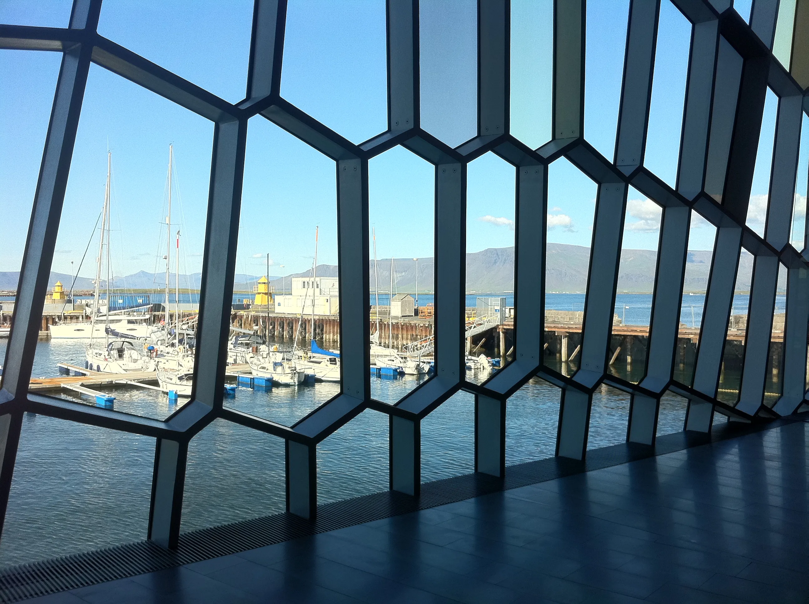 Vista desde el interior del Harpa Concert Hall a través de su fachada de cristal con forma de panal geométrico hacia el puerto con veleros y montañas al fondo