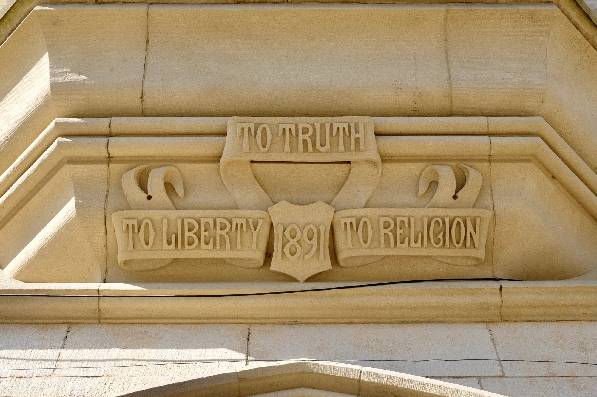 Gevelbeeldhouwwerk met een boekrol waarop de inscripties 'TO TRUTH', 'TO LIBERTY', 'TO RELIGION' en het jaartal '1891' staan.