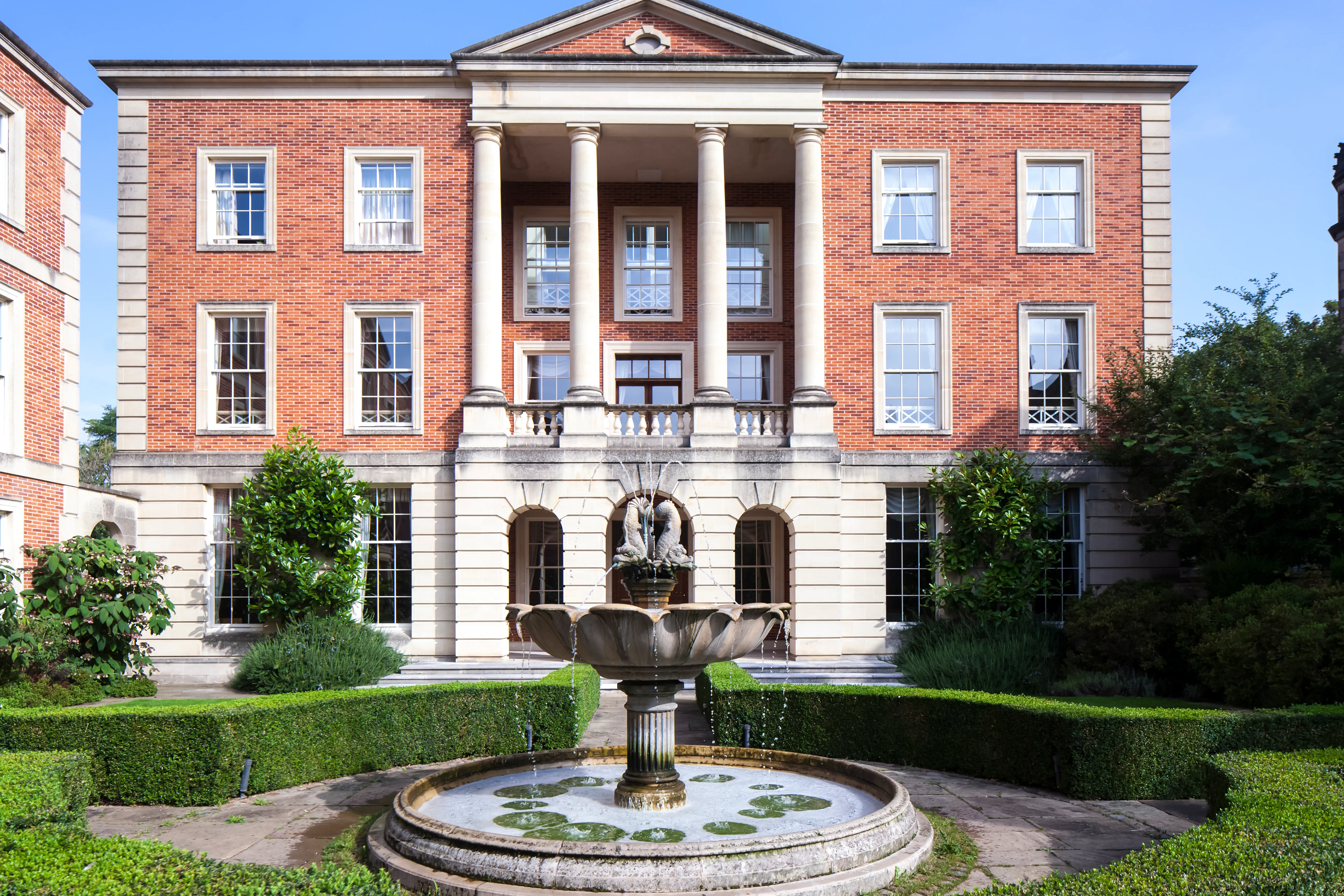 Neoclassicistisch gebouw van rode baksteen met witte stenen zuilen en een centrale portiek, met op de voorgrond een stenen fontein in een verzorgde tuin.