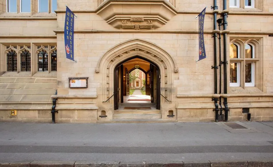 Stenen gevel van Harris Manchester College met een centrale boogvormige ingang die uitkijkt op een binnenplaats, geflankeerd door verticale blauwe banieren en ramen in gotische stijl.