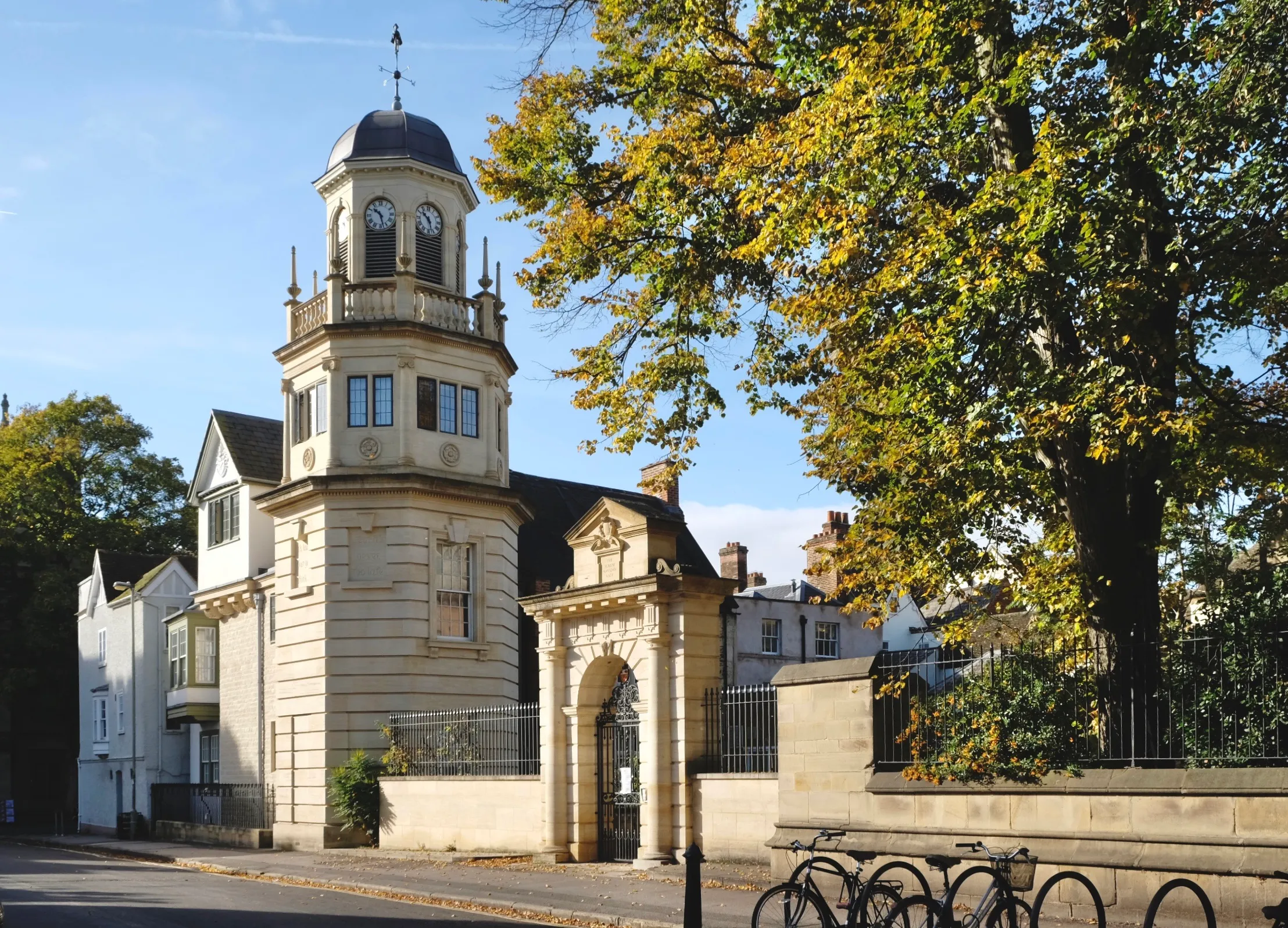 Buitenaanzicht van Harris Manchester College met de Siew-Sngiem Clock Tower met een koepelvormig torentje, stenen poortboog en herfstbomen met fietsen geparkeerd op de voorgrond