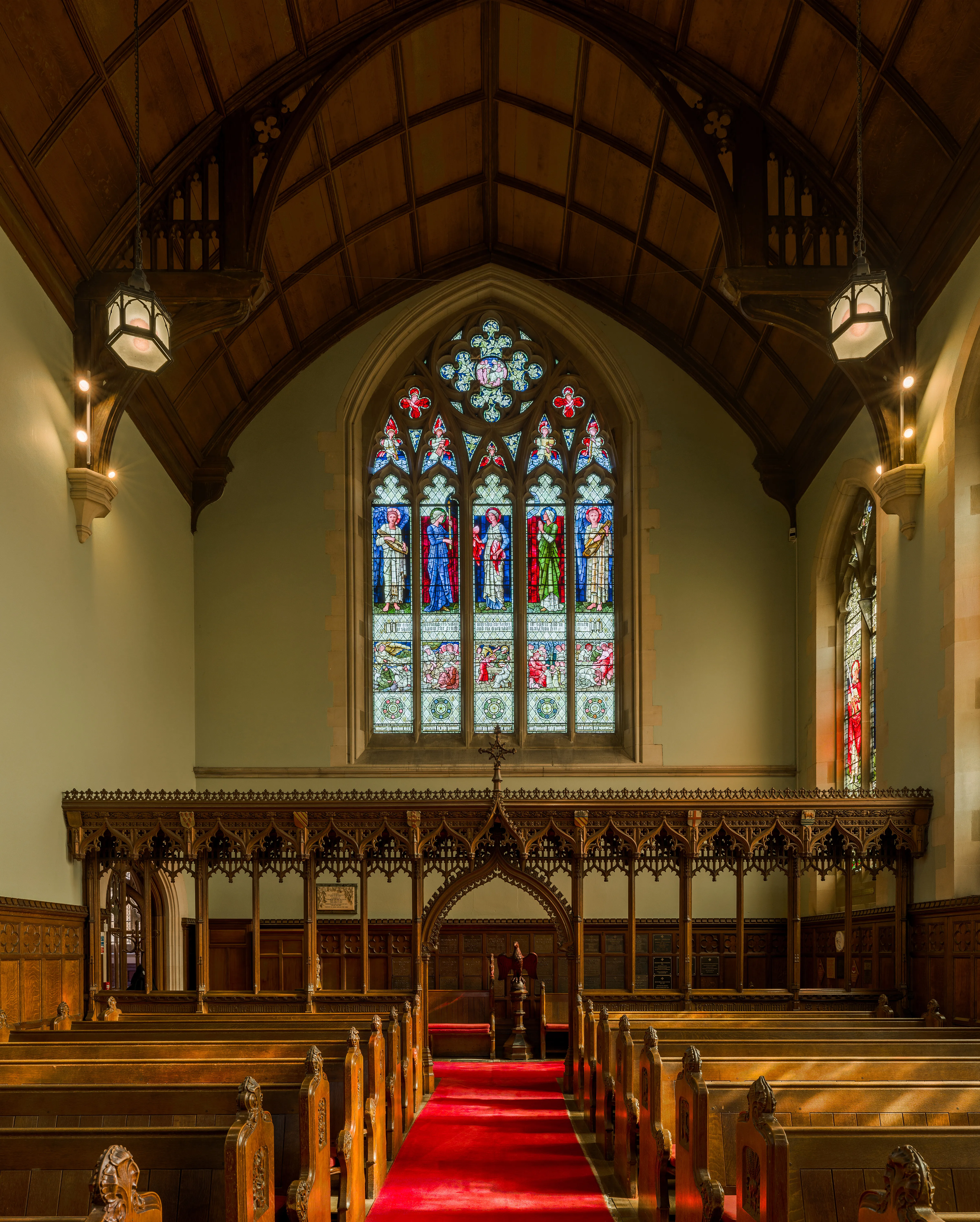 Interieur van de Harris Manchester College Chapel met houten kerkbanken, een rood tapijt in het gangpad, een rijkelijk versierd houten doksaal en een groot glas-in-loodraam onder een gewelfd houten plafond.