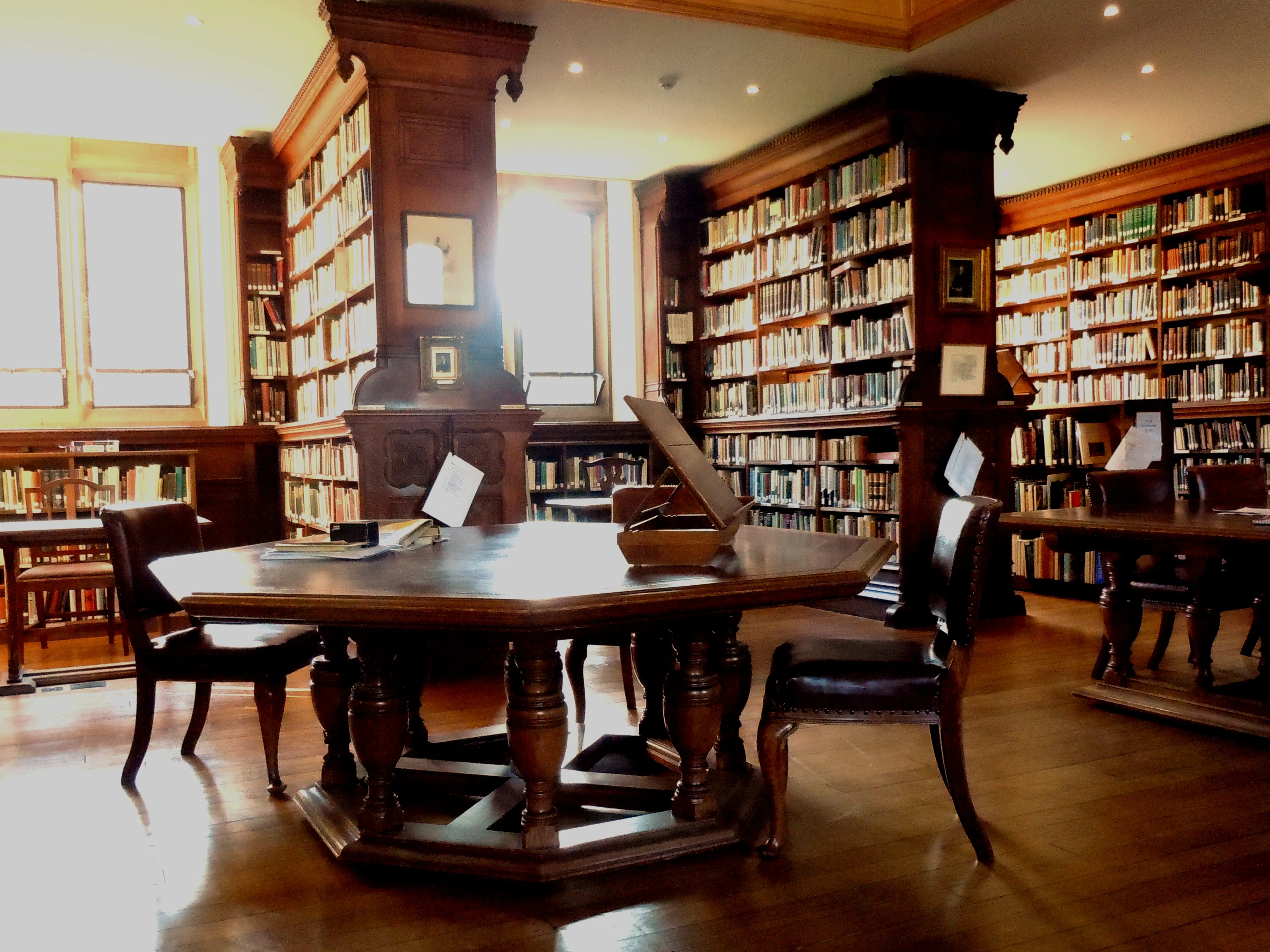 Interieur van een houten bibliotheek met kamerhoge boekenplanken vol boeken, een grote achthoekige leestafel omringd door stoelen en heldere ramen die de ruimte verlichten.