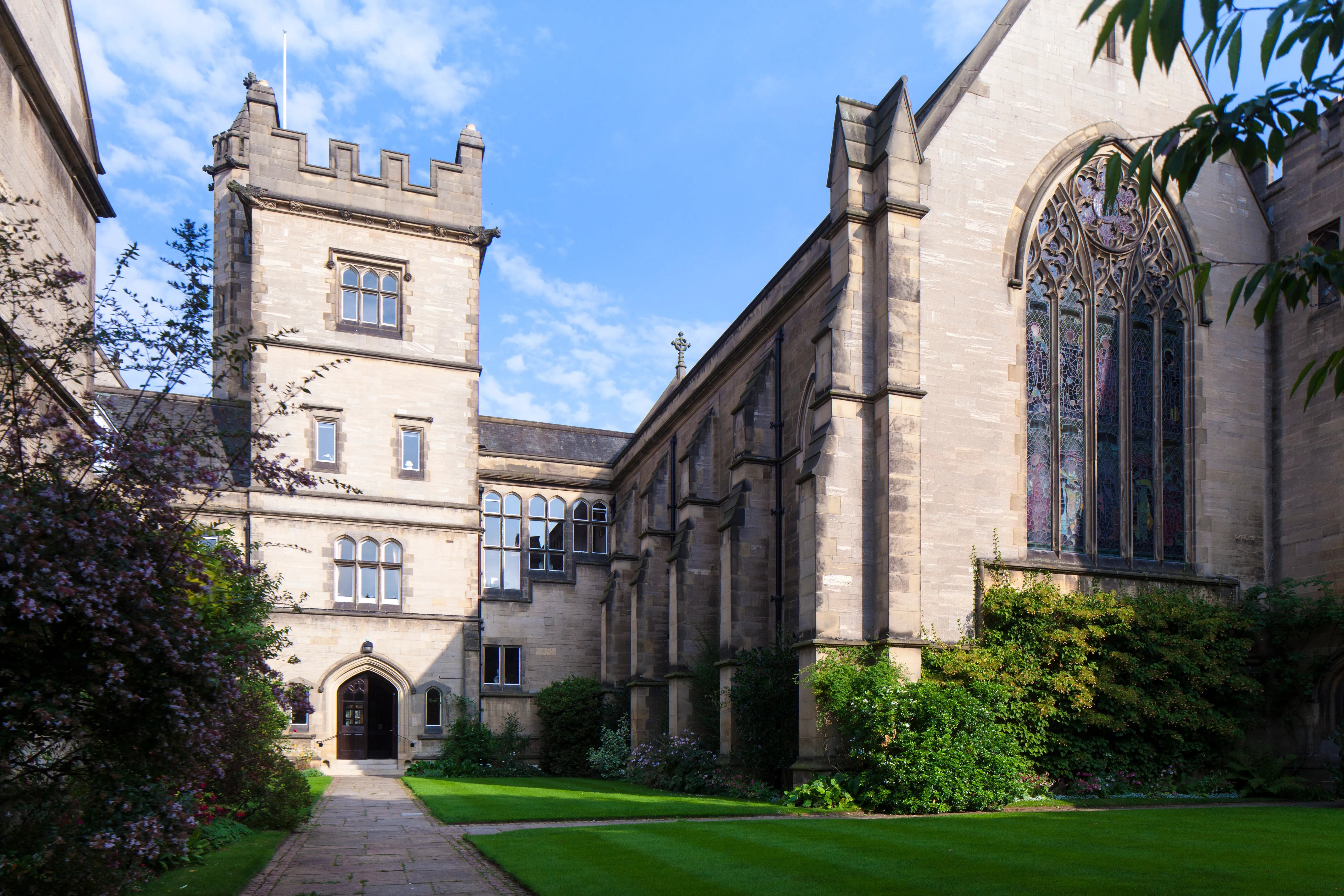 Stenen binnenplaats van Harris Manchester College met een gekanteelde toren, een gotisch kapelraam en een groen gazon onder een blauwe lucht