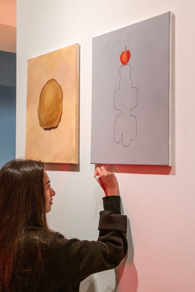 Woman adjusting two paintings on a gallery wall: one with brown shape on beige background, the other with red apple above abstract form on gray