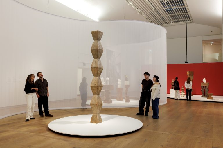 Modern art gallery with wooden geometric sculpture on circular platform, people observing, white walls and wooden floor