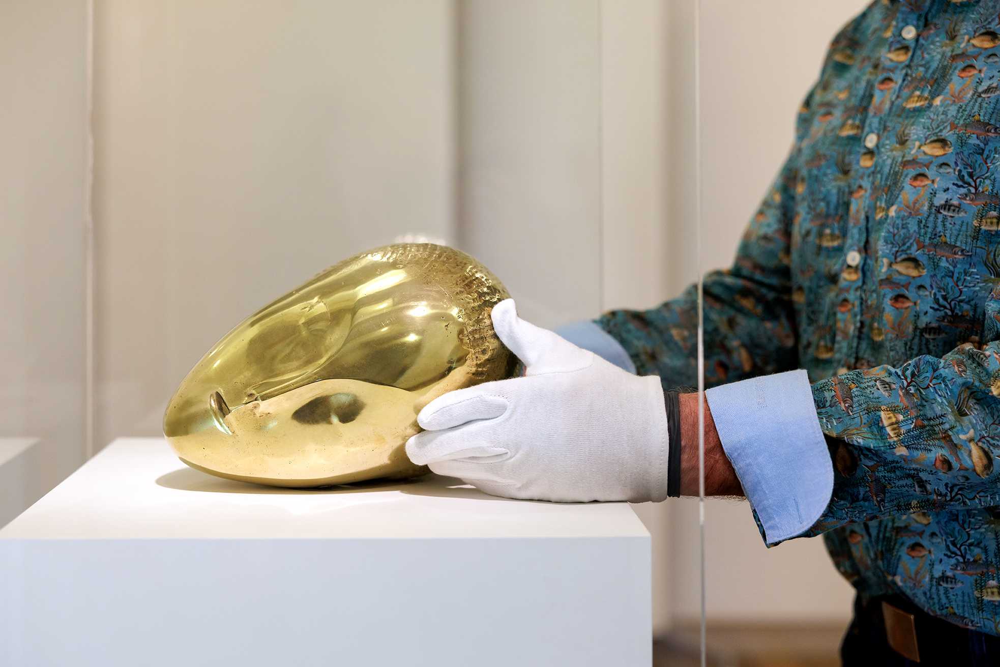 Person wearing white gloves handles a golden reflective sculptural artwork displayed on a white pedestal in a museum gallery