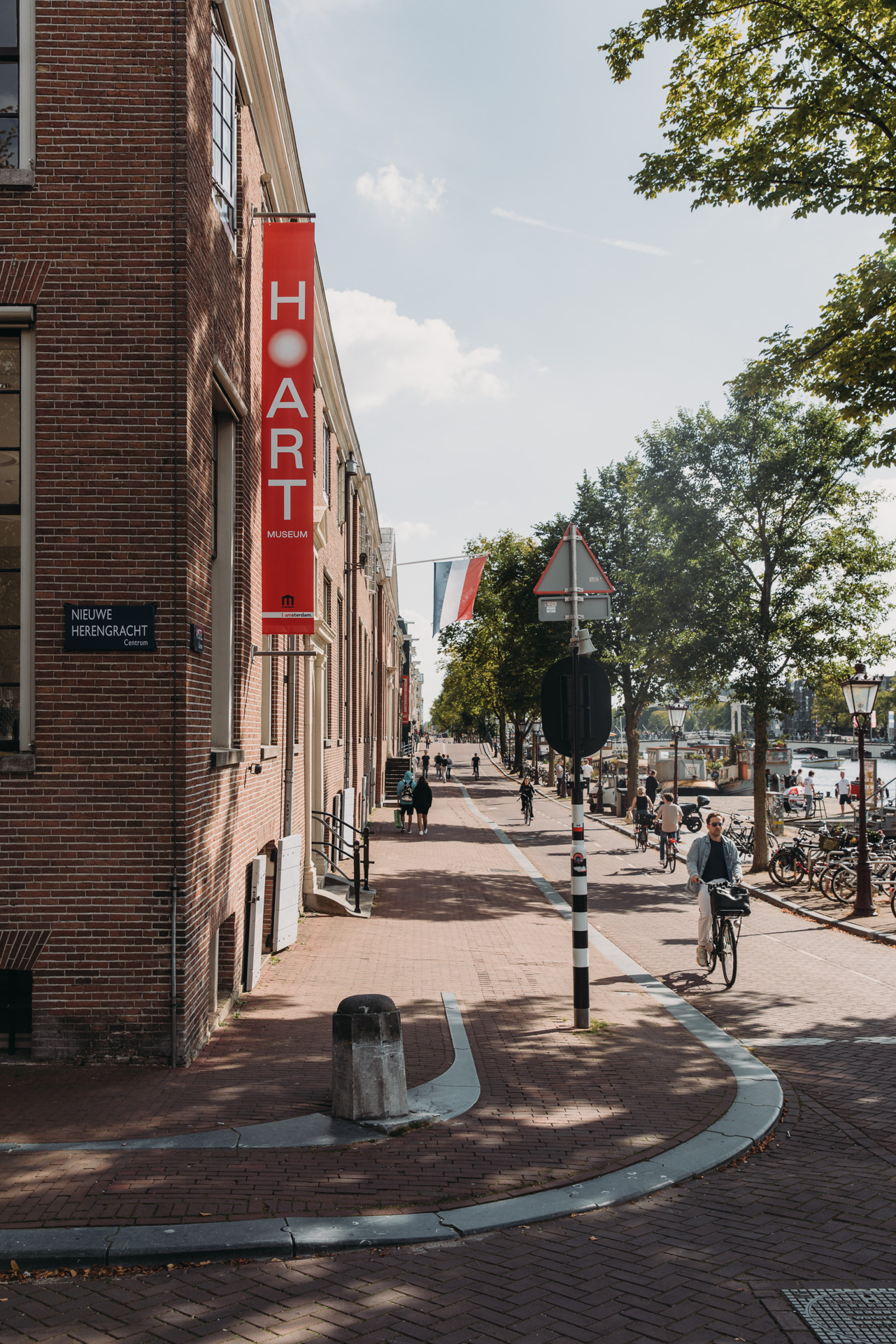 H'ART Museum building exterior with red vertical banner, brick facade, pedestrians, and cyclists on a tree-lined street