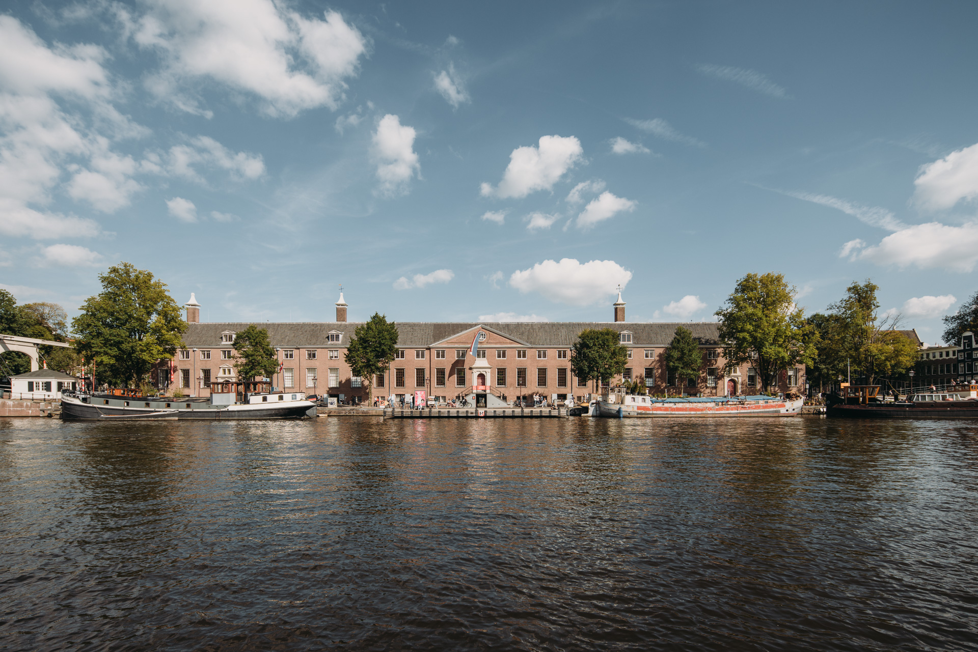 Long brick building with multiple chimneys along a river, boats docked, trees, and scattered clouds