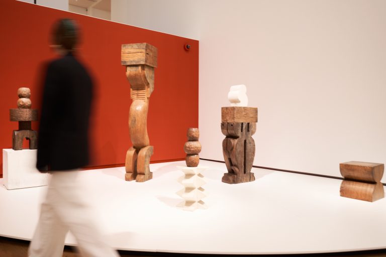 Wooden sculptures arranged on white platforms against a red wall in an exhibition space with a blurred figure walking by