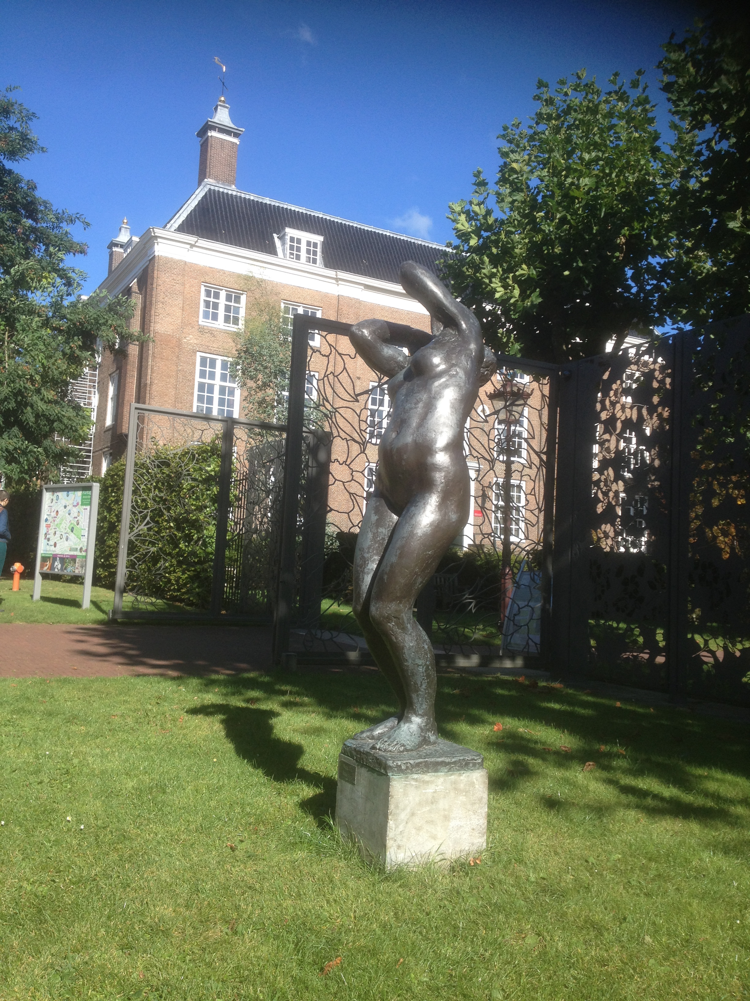 Bronze sculpture of a figure on a pedestal in front of a brick building with a black roof and white windows