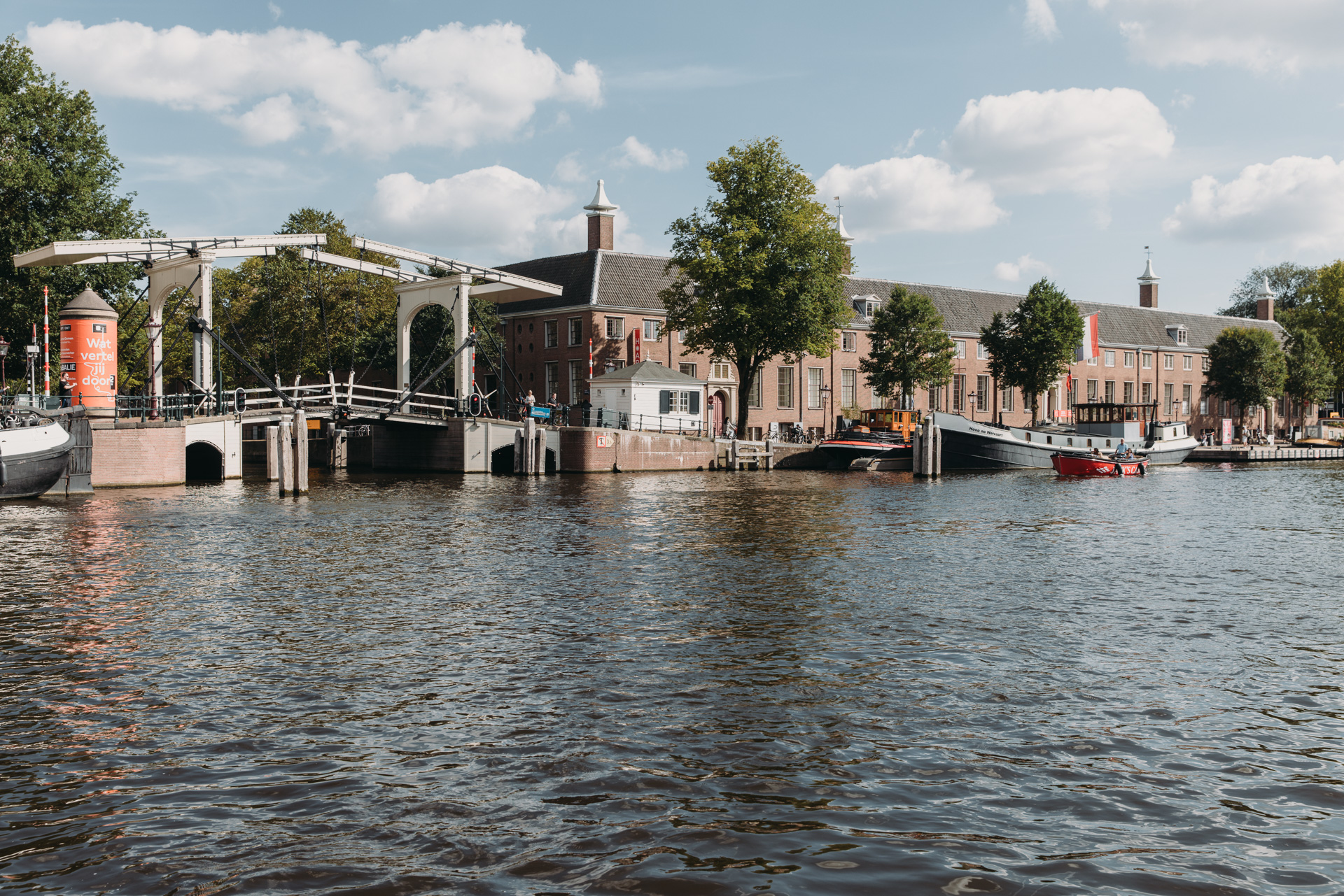 Historic brick building with gabled roof beside canal, white pedestrian bridge with people, blue sky with clouds, boats on water