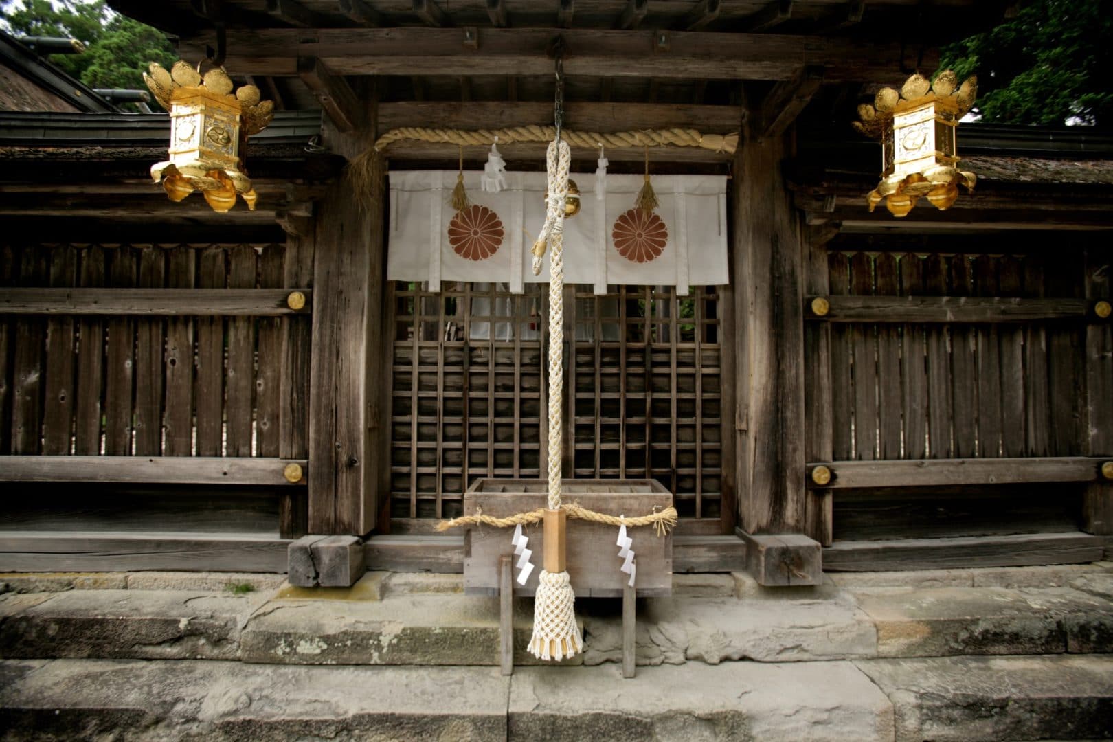 Traditional Japanese shrine entrance with wooden lattice doors, stone steps, golden lanterns, and sacred shimenawa rope