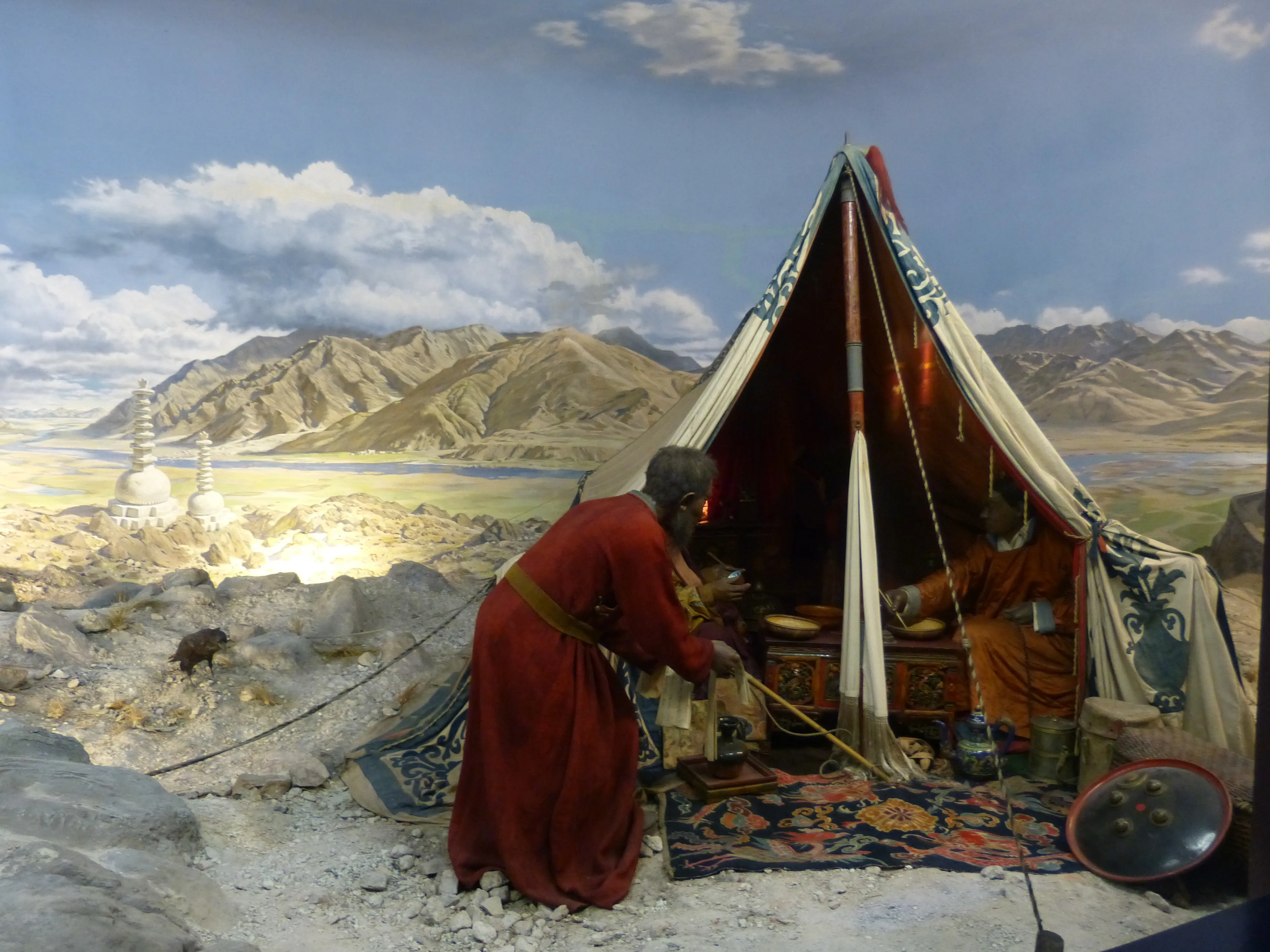 Diorama de museo que representa una escena de un campamento tibetano con figuras vestidas con trajes tradicionales junto a una tienda decorada, sobre un telón de fondo de paisaje montañoso pintado con estupas blancas.