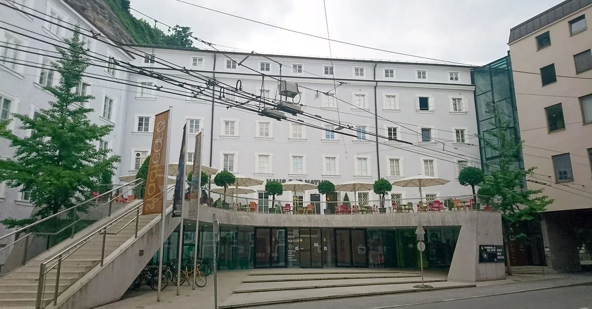 Fachada de un edificio blanco de varias plantas con una terraza exterior con sombrillas, escaleras de piedra que conducen a la entrada y una entrada acristalada a nivel de calle con cables de tranvía por encima.