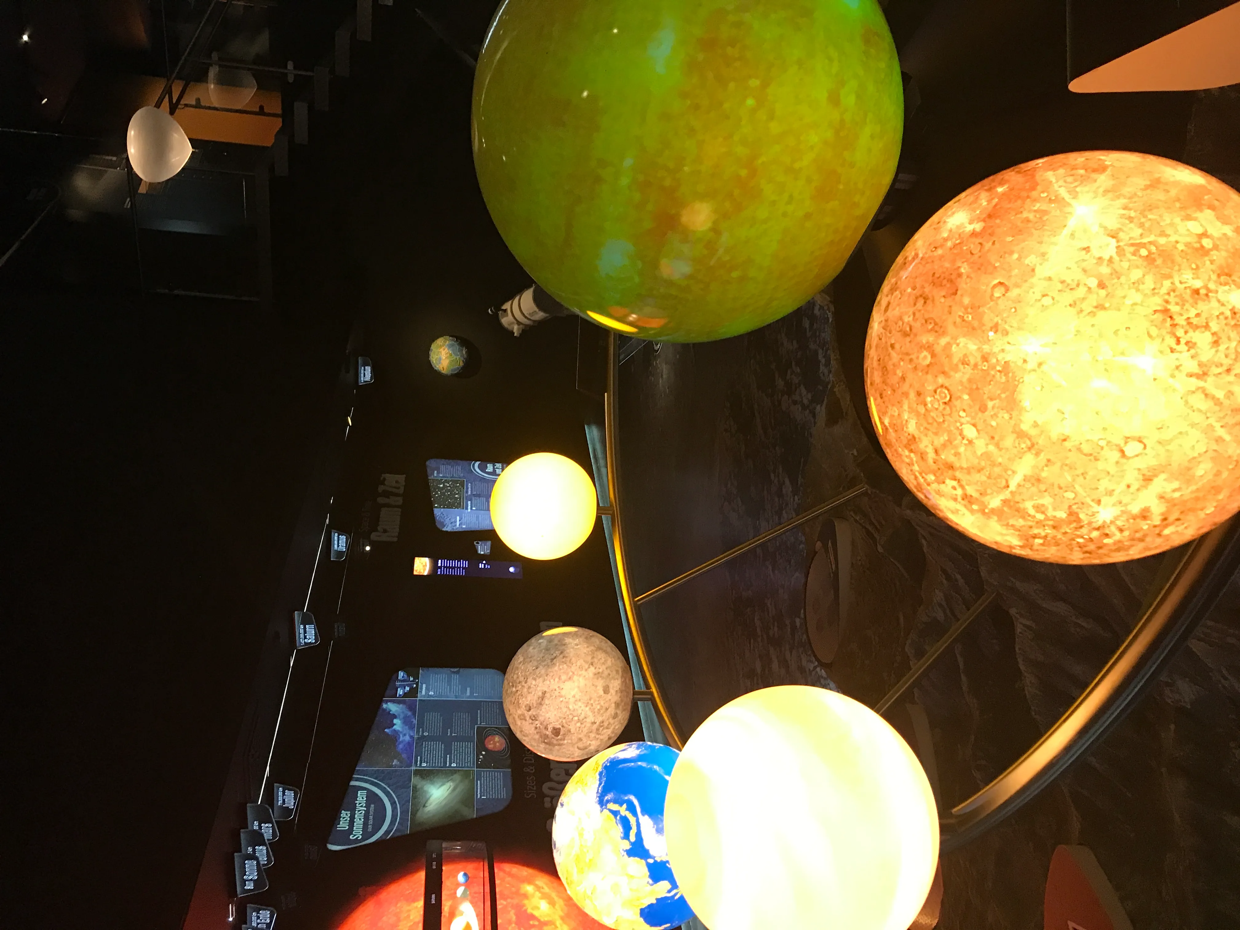 Modelos de planetas brillantes e iluminados de varios tamaños y colores que cuelgan de un techo oscuro en el espacio de exposición de un museo, incluyendo un gran planeta verde, una esfera naranja similar al sol y un modelo de la Tierra.