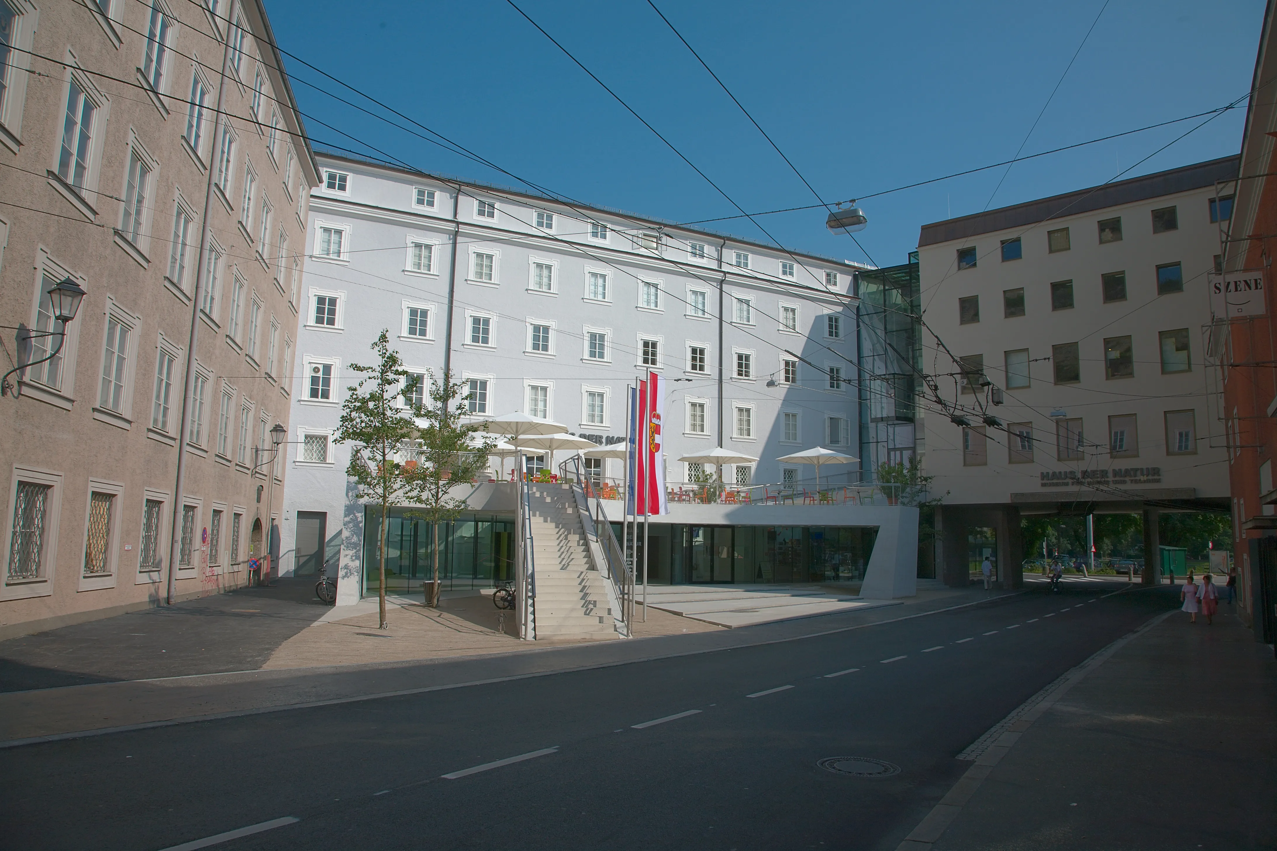 Vista de calle del museo Haus der Natur en Salzburgo, que muestra una moderna estructura de entrada de cristal situada entre edificios históricos con un estandarte rojo colgado de la fachada.
