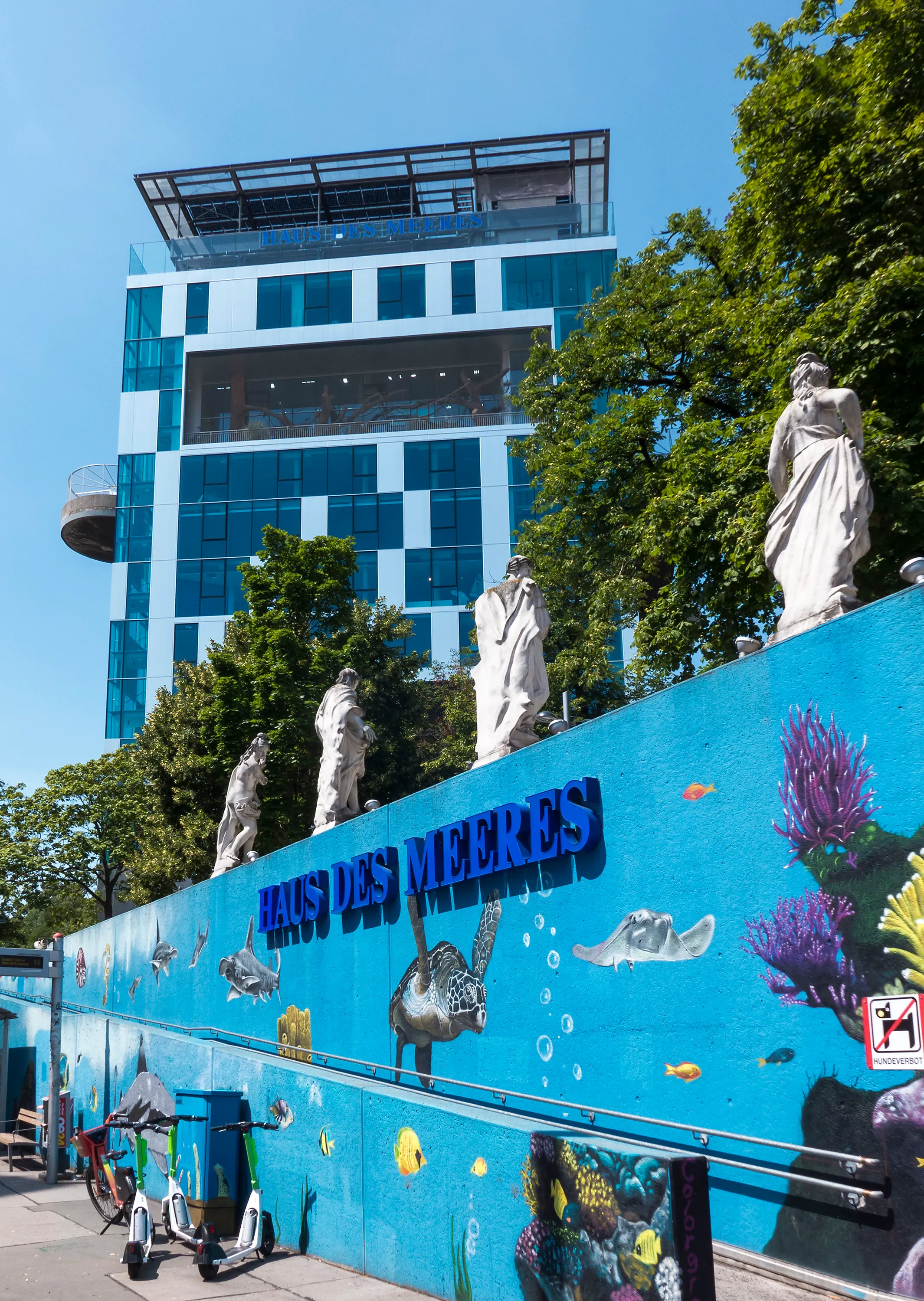 Blauwe muur met maritieme muurschilderingen en witte beelden voor een modern glazen gebouw met de tekst 'HAUS DES MEERES'