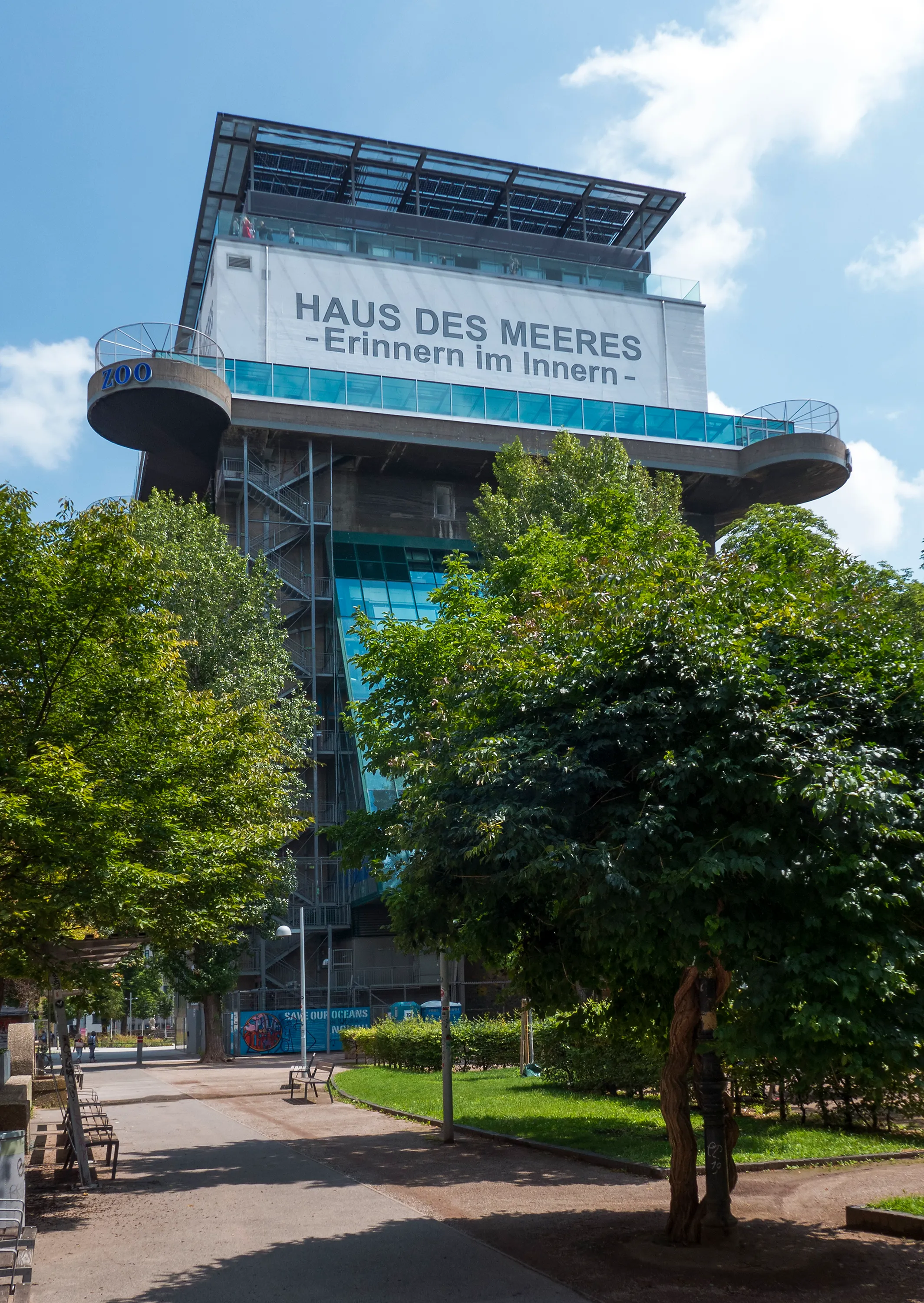 Een grote betonnen flakkenkoepel met een bord met de tekst 'HAUS DES MEERES -Erinnern im Innern-', omringd door groene bomen en een verhard pad onder een blauwe lucht.