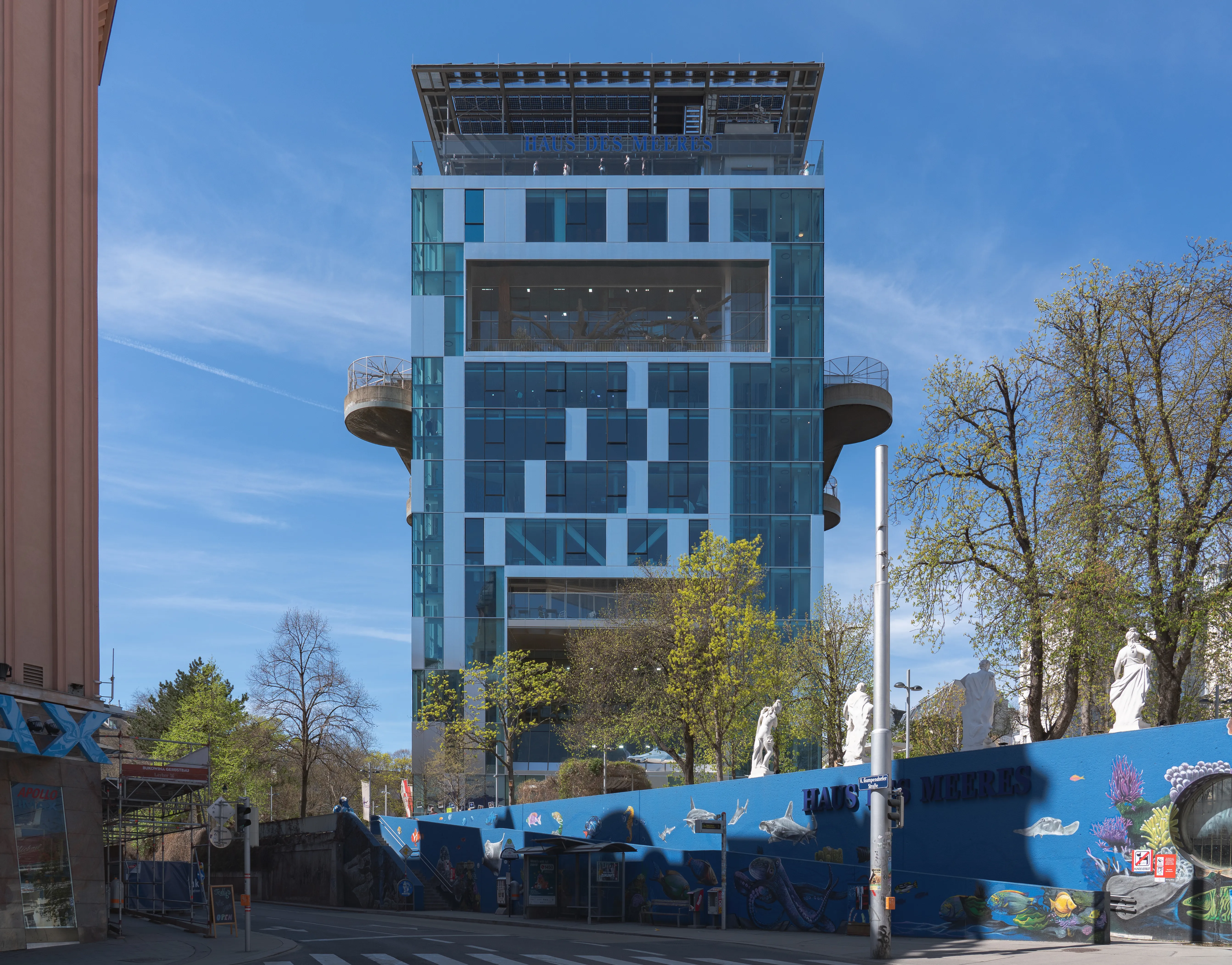 Hoge toren van glas en beton met cirkelvormige observatiedekken, omgeven door bomen en een blauwe muur met kunstwerken