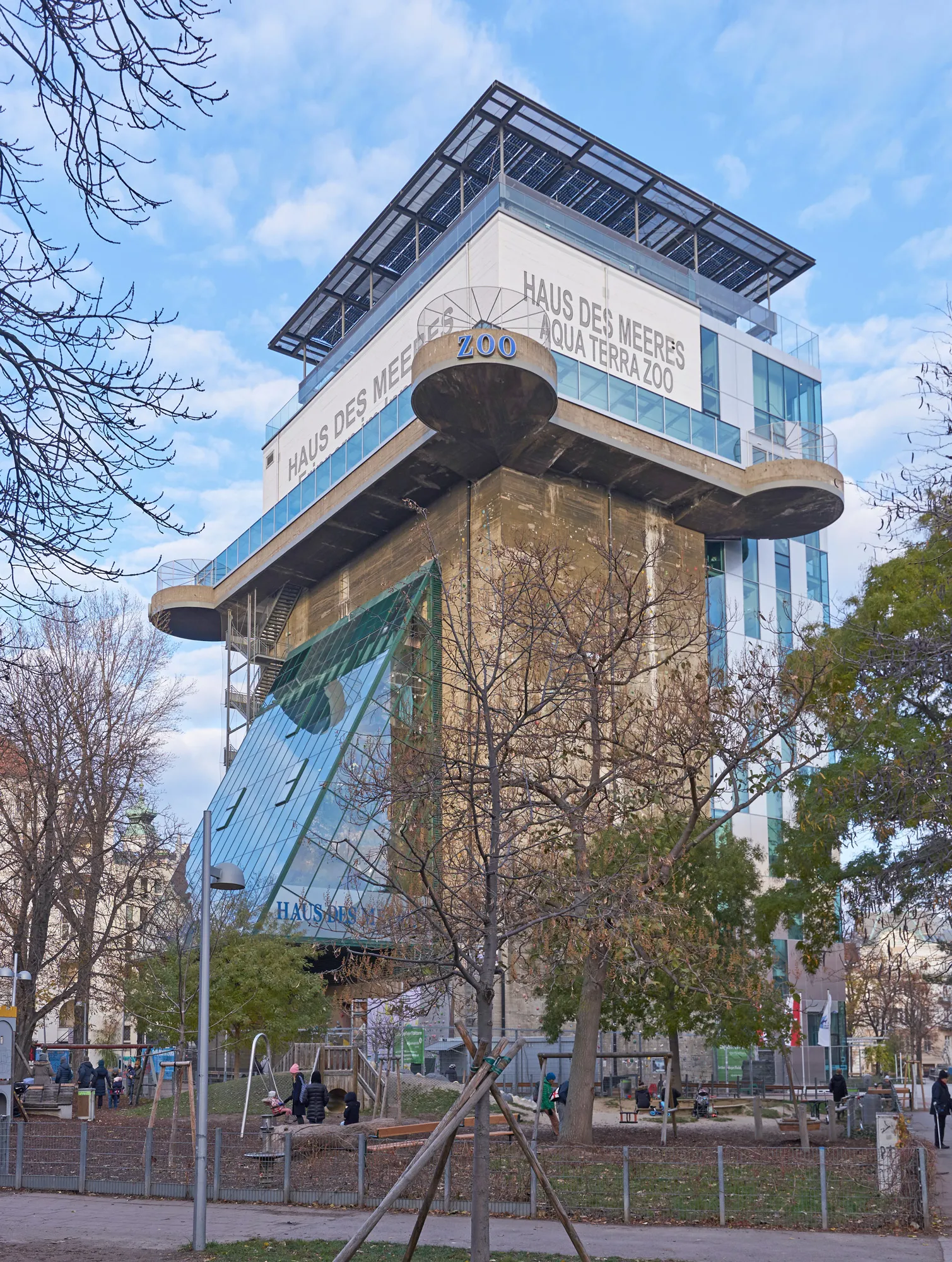 Een grote betonnen flak toren met moderne glazen uitbreidingen en een bord met de tekst 'HAUS DES MEERES AQUA TERRA ZOO' aan de zijkant, omringd door bomen en een parkgebied.