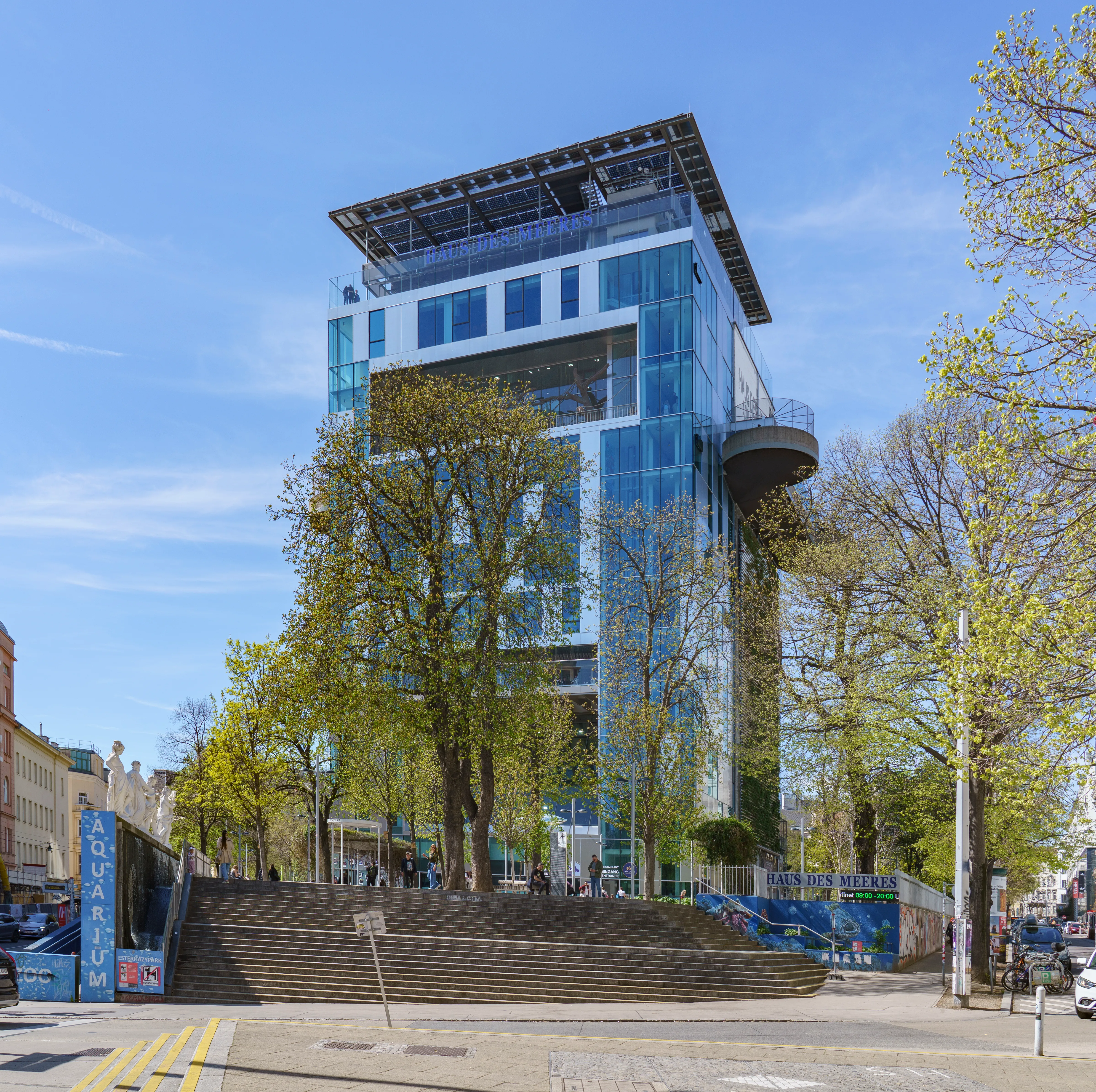 Een modern glazen gebouw met een kenmerkende bovenstructuur, omgeven door bomen en een brede trap die naar de ingang leidt.