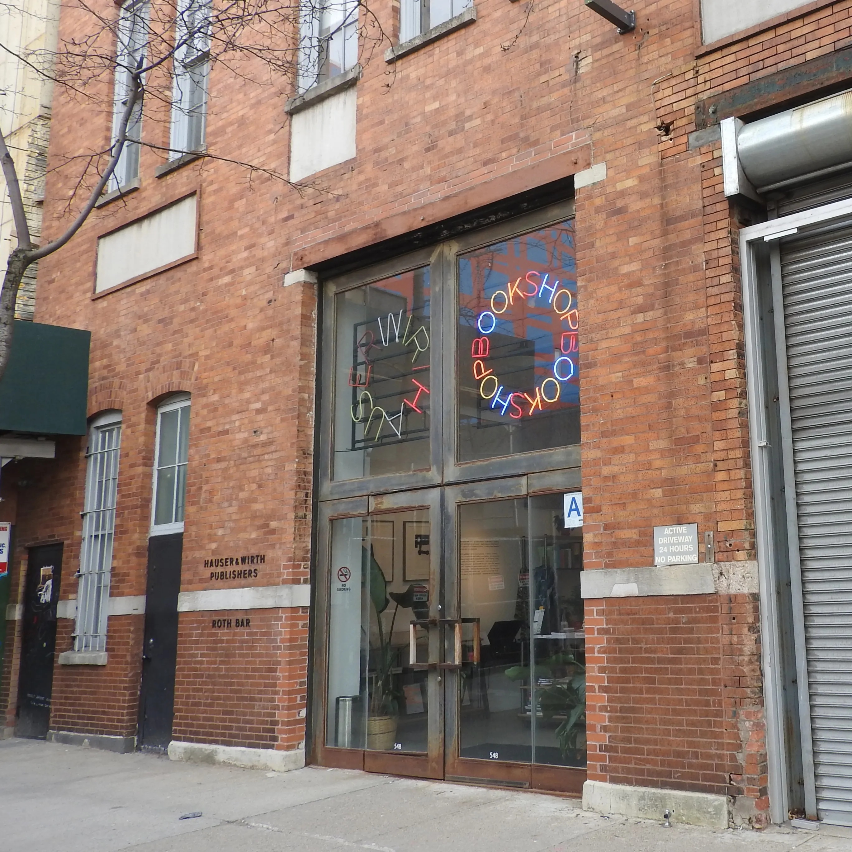 Vista exterior de la galería Hauser & Wirth en un edificio de ladrillo rojo, que cuenta con una fachada de cristal con un letrero de neón circular que dice 'BOOKSHOP' y el texto 'H&W' en el escaparate.