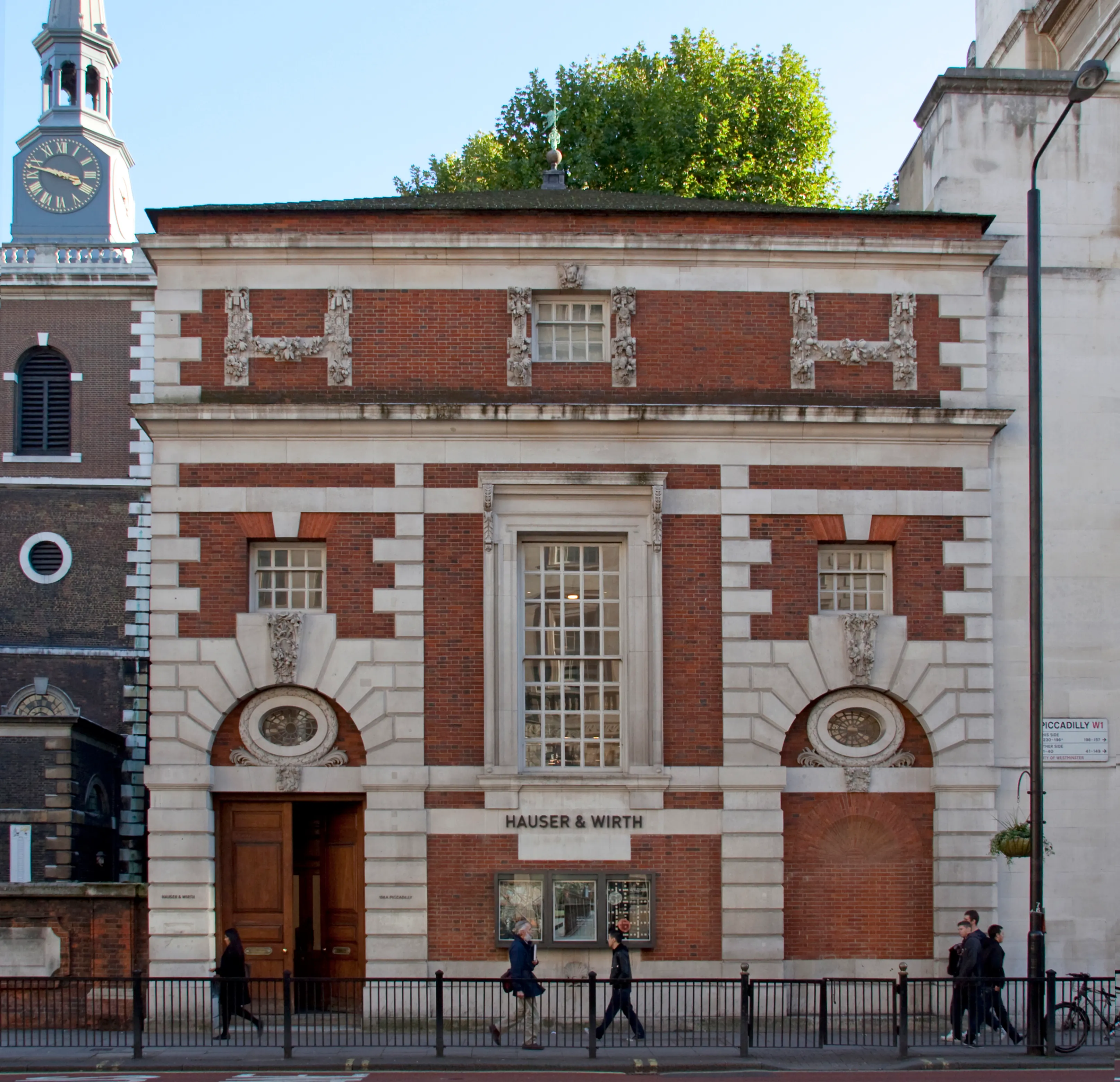 Fachada de un edificio neoclásico de ladrillo rojo con detalles en piedra blanca y el rótulo de Hauser & Wirth sobre la entrada, con la torre de un reloj de iglesia visible a la izquierda.