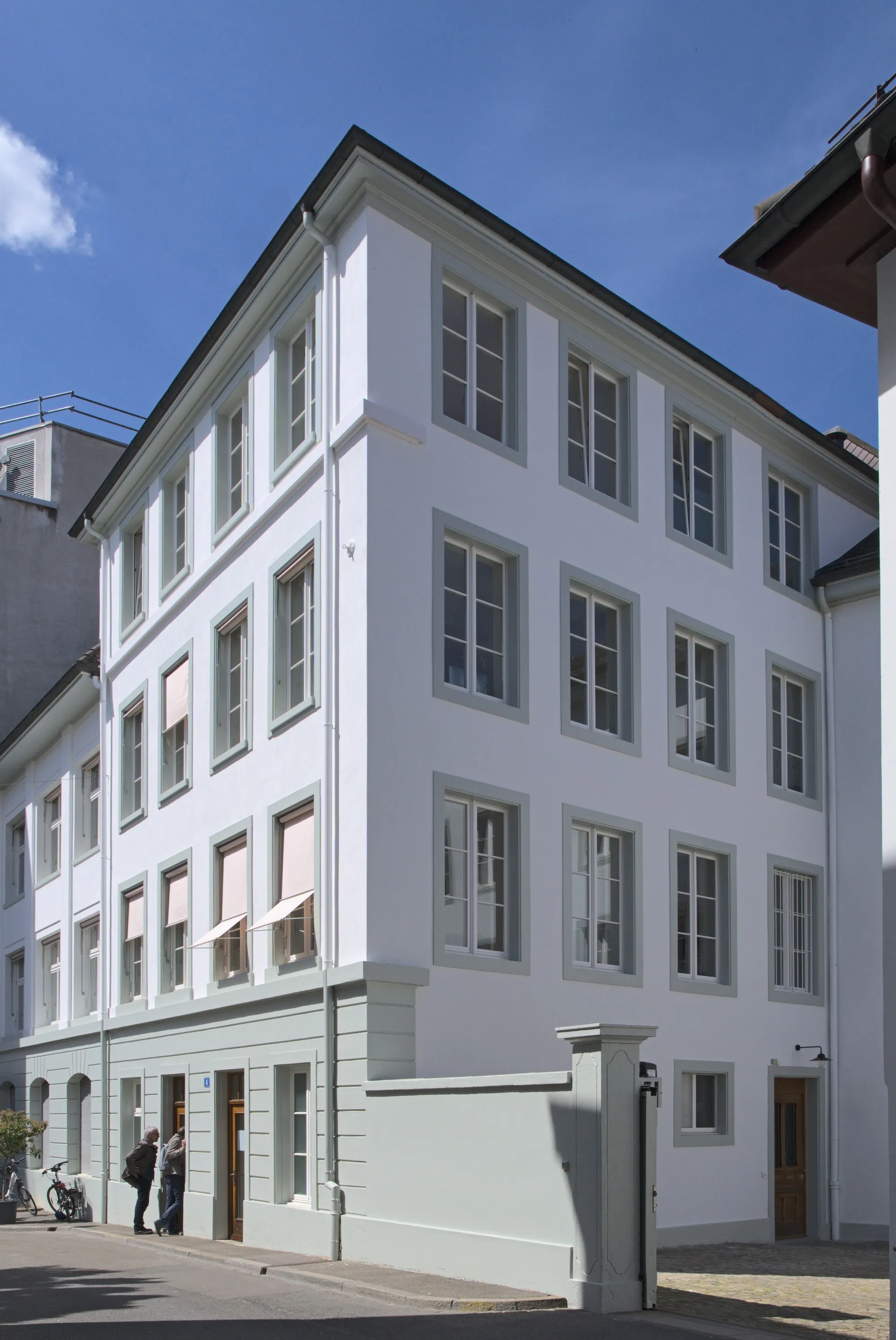 Edificio blanco de varias plantas con marcos de ventana grises y toldos rosas; la galería Hauser & Wirth Basel en el casco antiguo, con personas cerca de la entrada bajo un cielo azul.