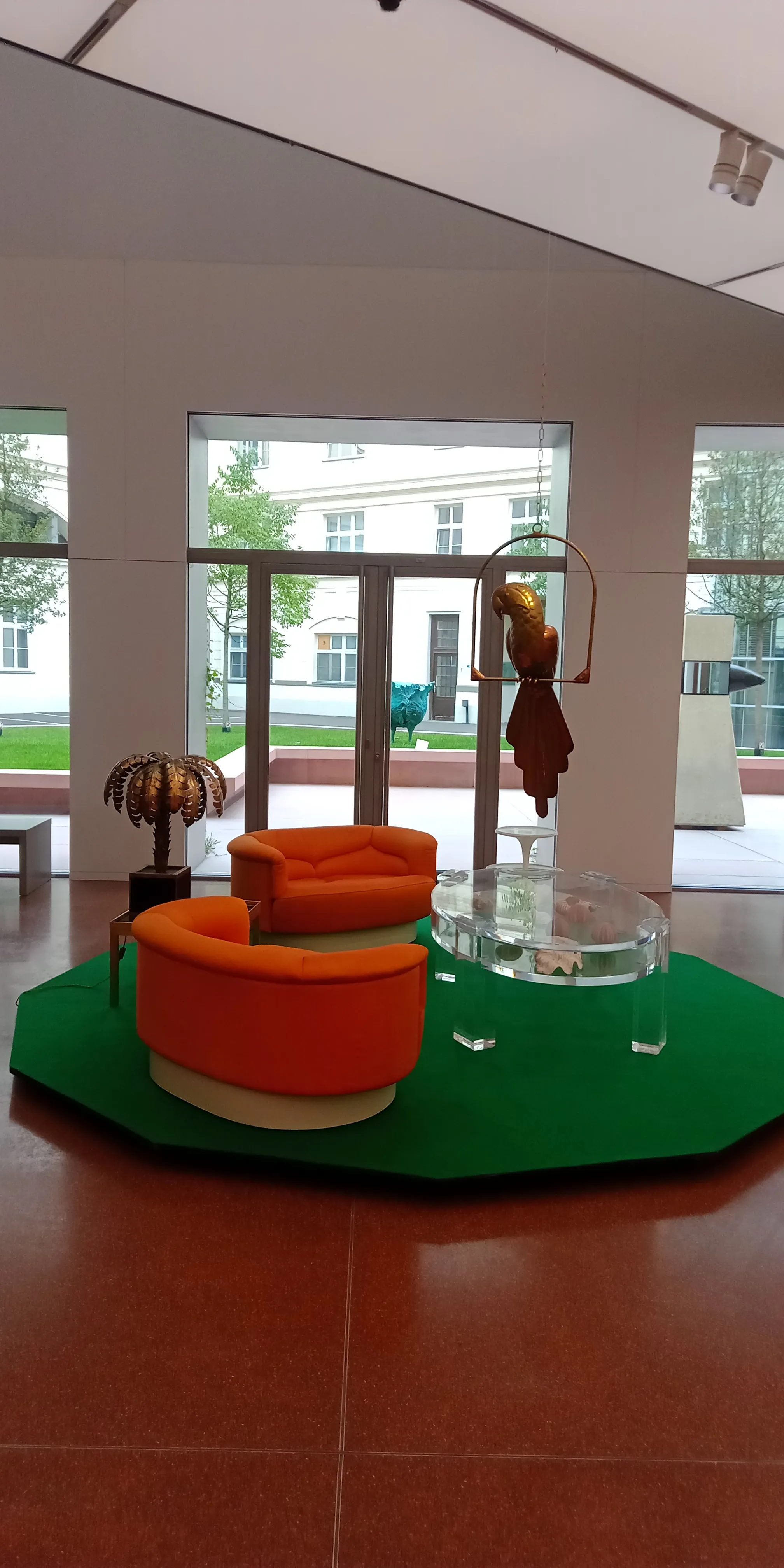 Modern interior space with orange sofas, a glass table, and a hanging sculpture on a green platform