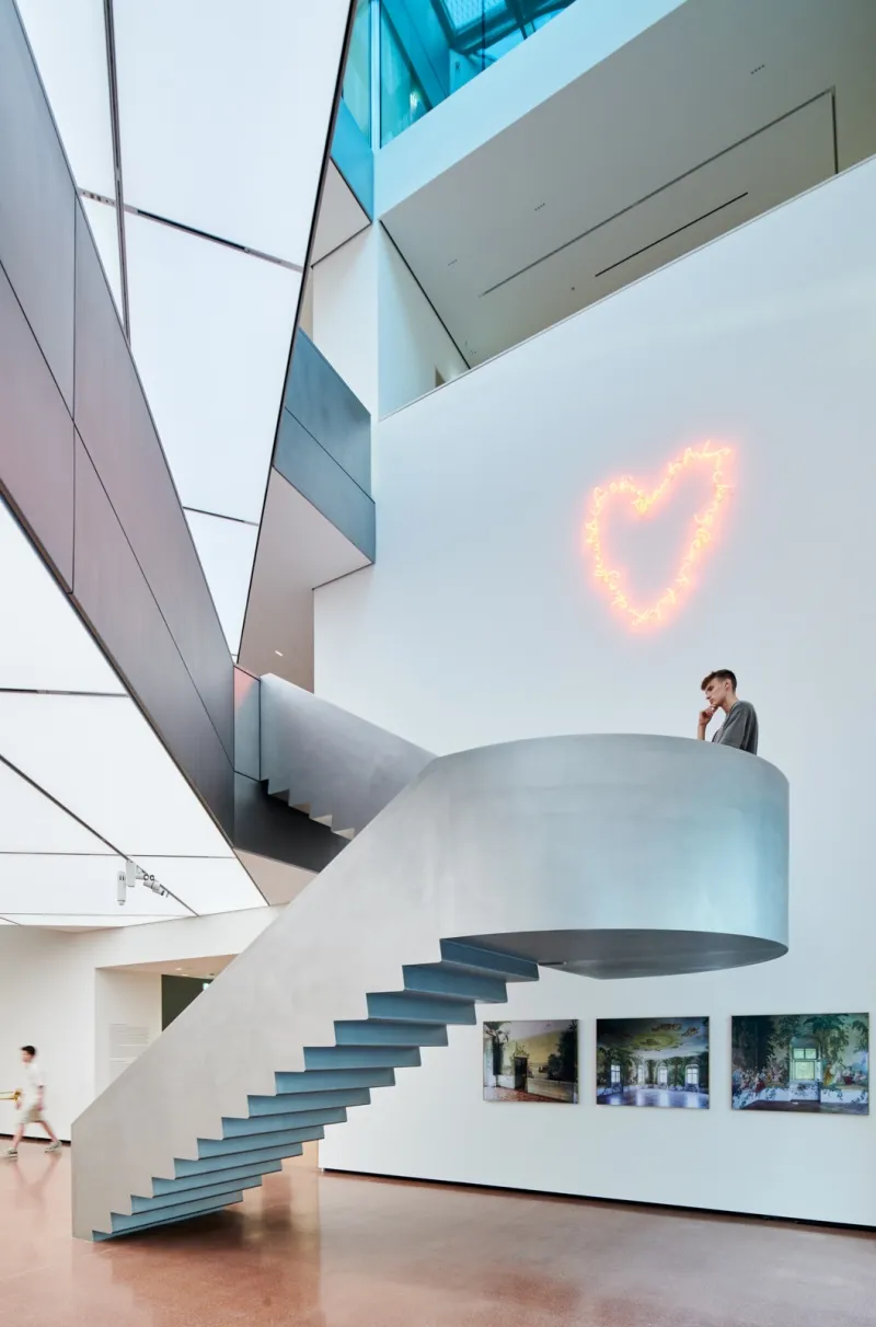 A curved staircase in a modern art museum with a glowing heart-shaped light installation on the wall and framed artworks below