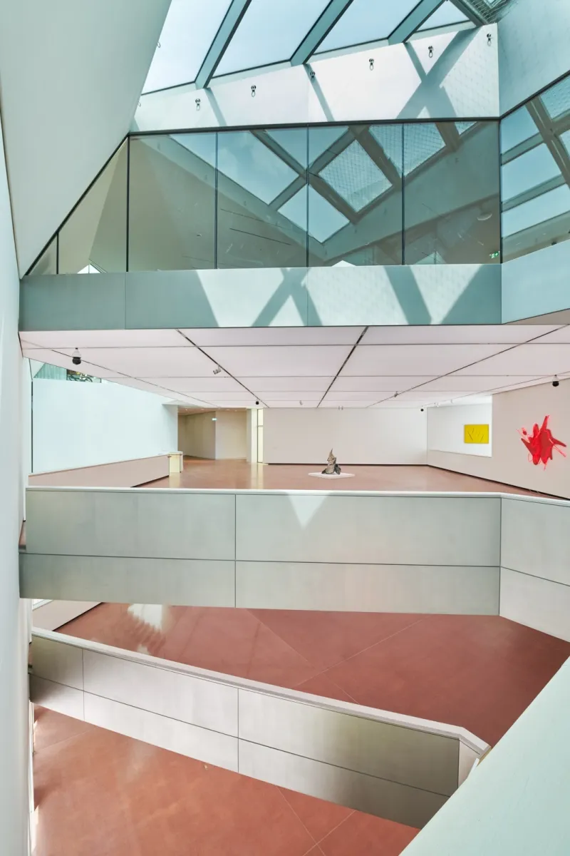 Modern interior atrium with glass ceiling, white walls, and terracotta floors, featuring a central sculpture and a red artwork on the wall