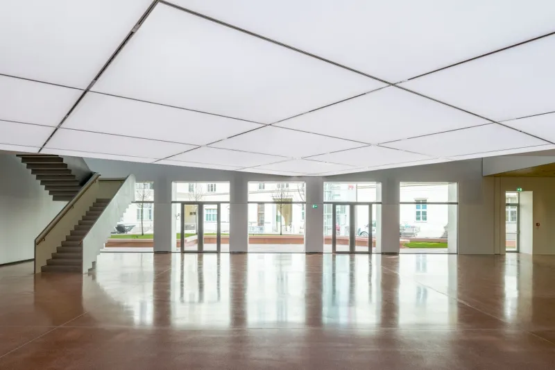 Spacious interior hall with a geometric ceiling grid, polished wooden floor, and large windows