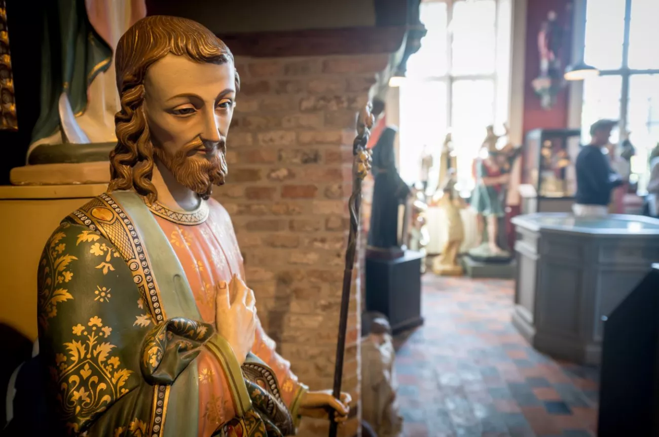 Estatua de madera pintada de un santo con túnicas ornamentadas con patrones dorados sosteniendo un bastón, exhibida dentro del Heiligenmuseum junto a otras estatuas y un visitante visible en el fondo borroso.