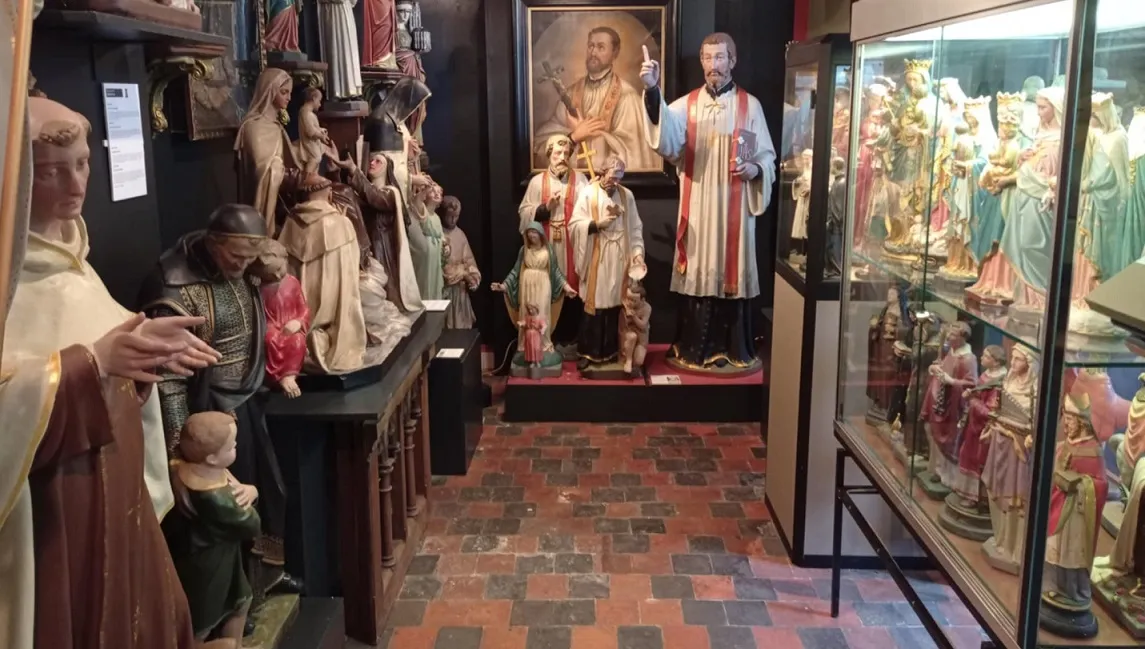 Interior de un museo lleno de coloridas estatuas religiosas de santos expuestas en estantes de madera y vitrinas, con un gran retrato pintado en la pared del fondo y suelo de ladrillo rojo