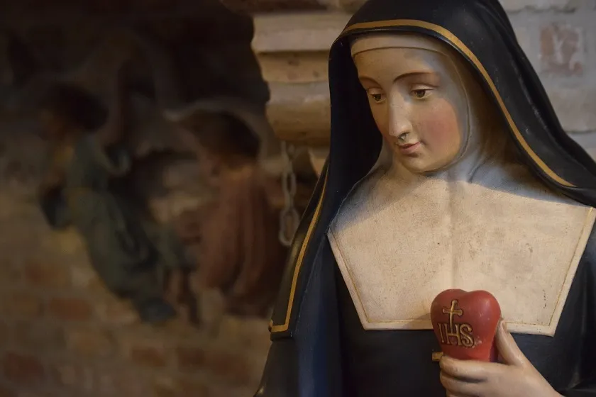 Primer plano de una estatua de madera pintada de una monja con hábito azul oscuro que sostiene un corazón rojo con la inscripción IHS