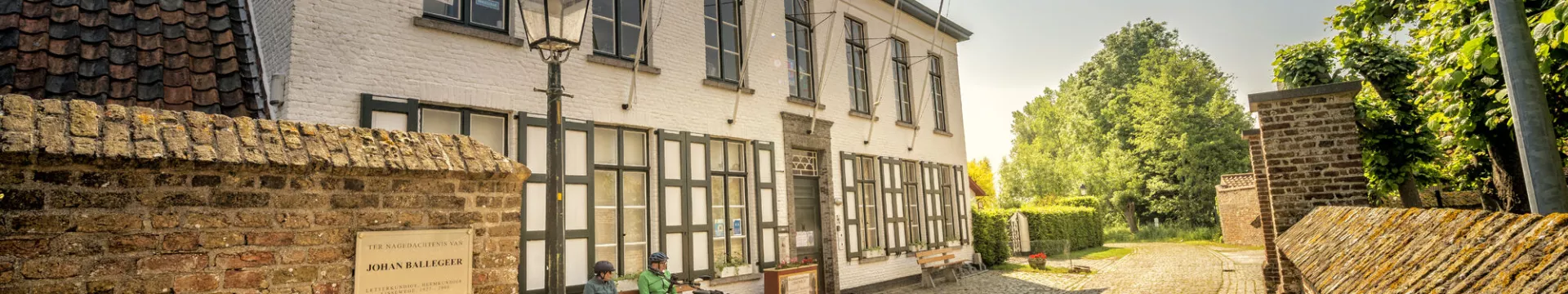 Edificio de yeso blanco con ventanas de marco oscuro detrás de un muro de ladrillo, con una farola y árboles verdes visibles, exterior del Heiligenmuseum en Lissewege