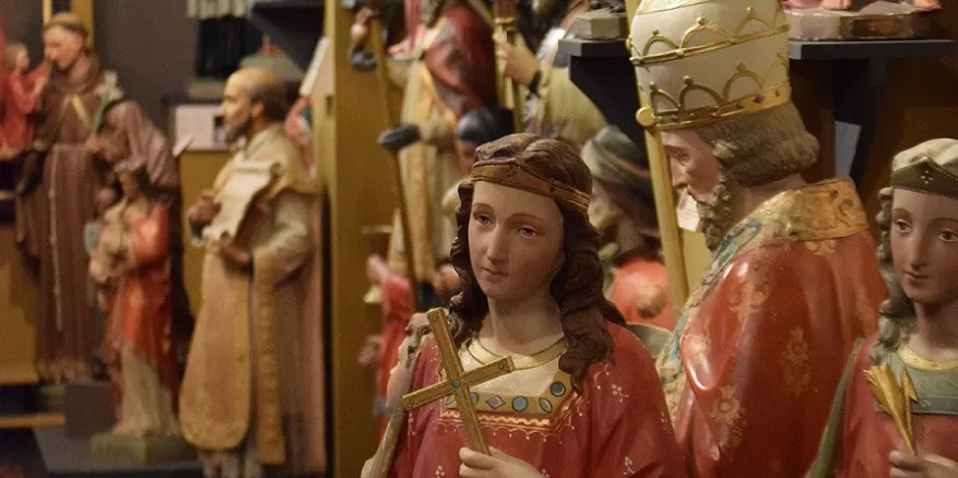 Estatuas de madera pintada de santos con túnicas coloridas, con una figura central que sostiene una cruz, expuestas en un museo.